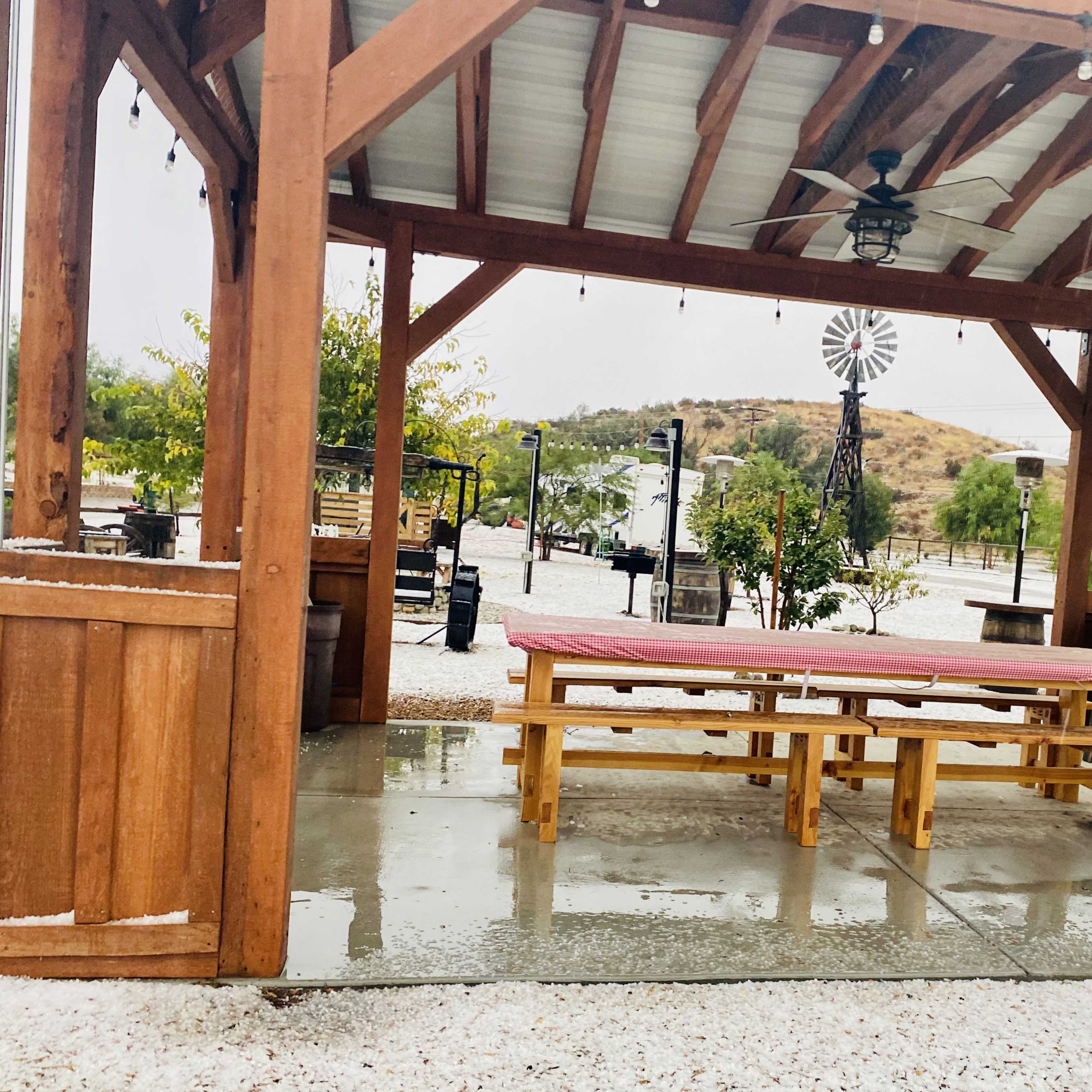 A covered outdoor patio features wooden picnic tables and a ceiling fan, set against a backdrop of rolling hills and a windmill.