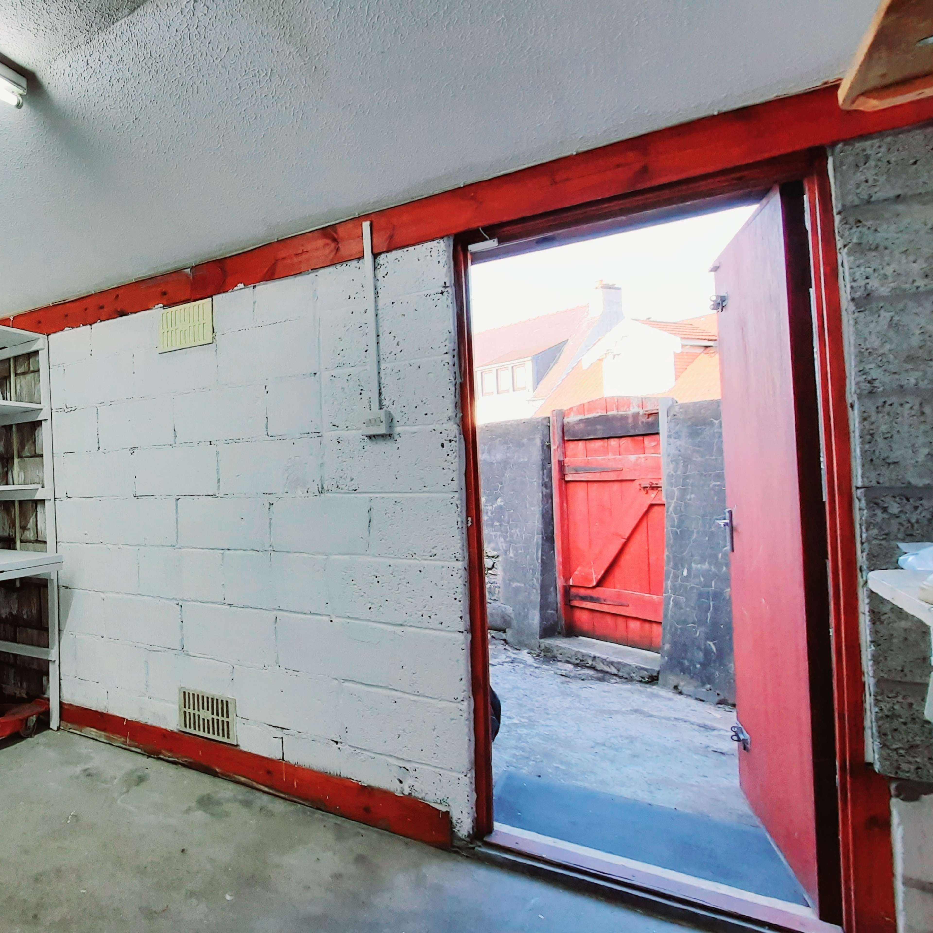A small, empty room with shelving on one side and an open doorway leading to an outdoor area with a red gate.