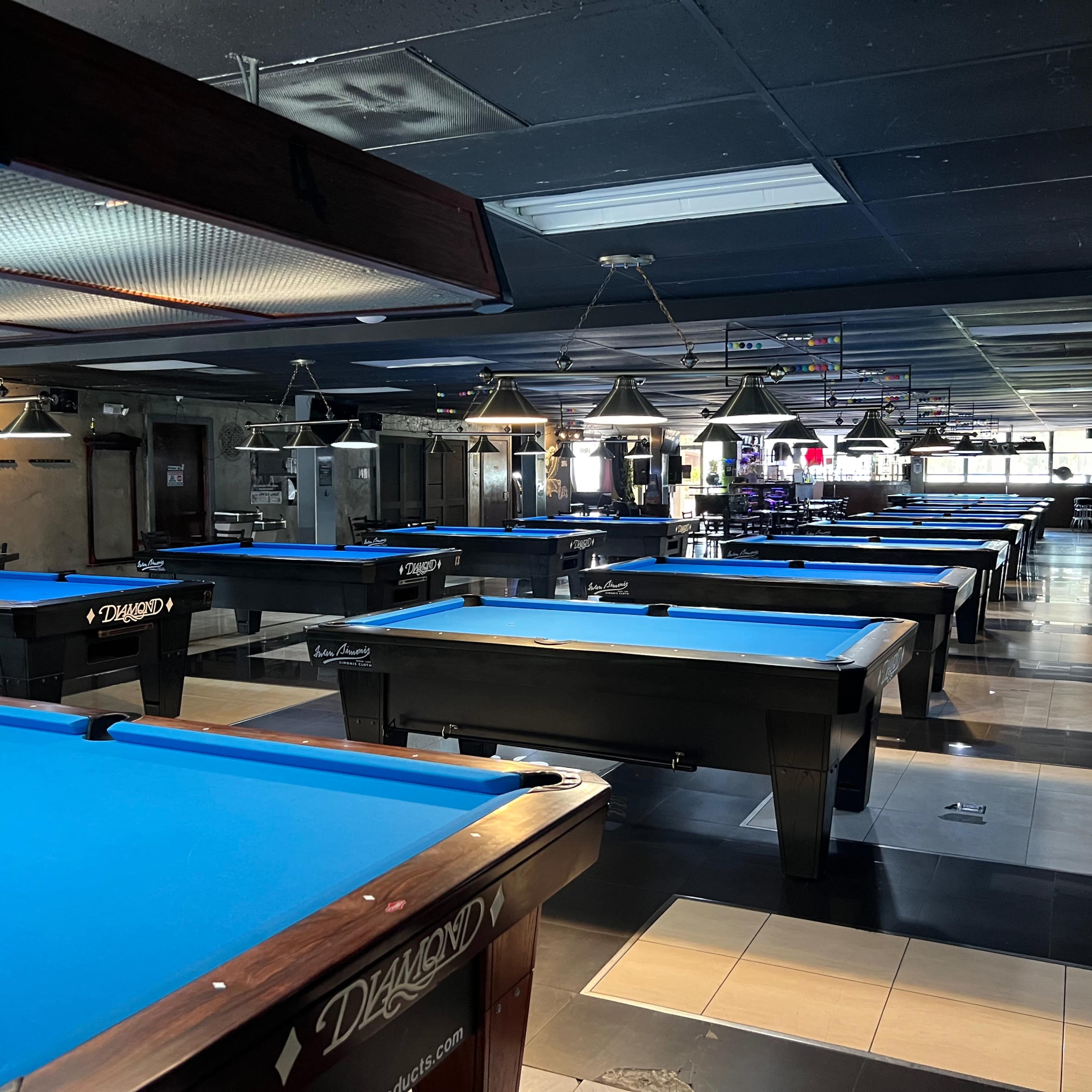 A spacious billiards hall filled with several blue-felt pool tables arranged in rows under bright overhead lighting.