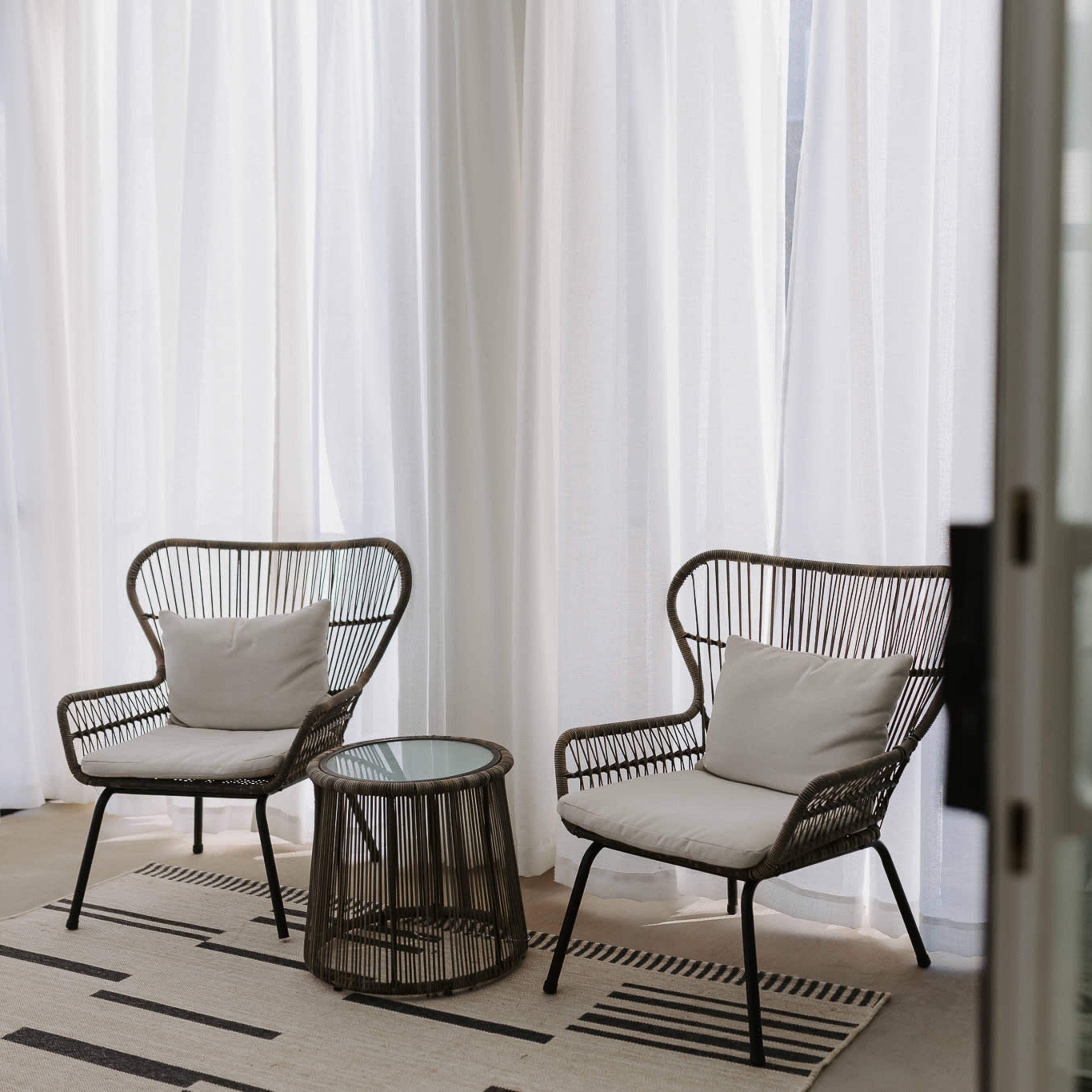 A cozy seating area featuring two wicker chairs with cushions and a glass side table, surrounded by sheer white curtains.