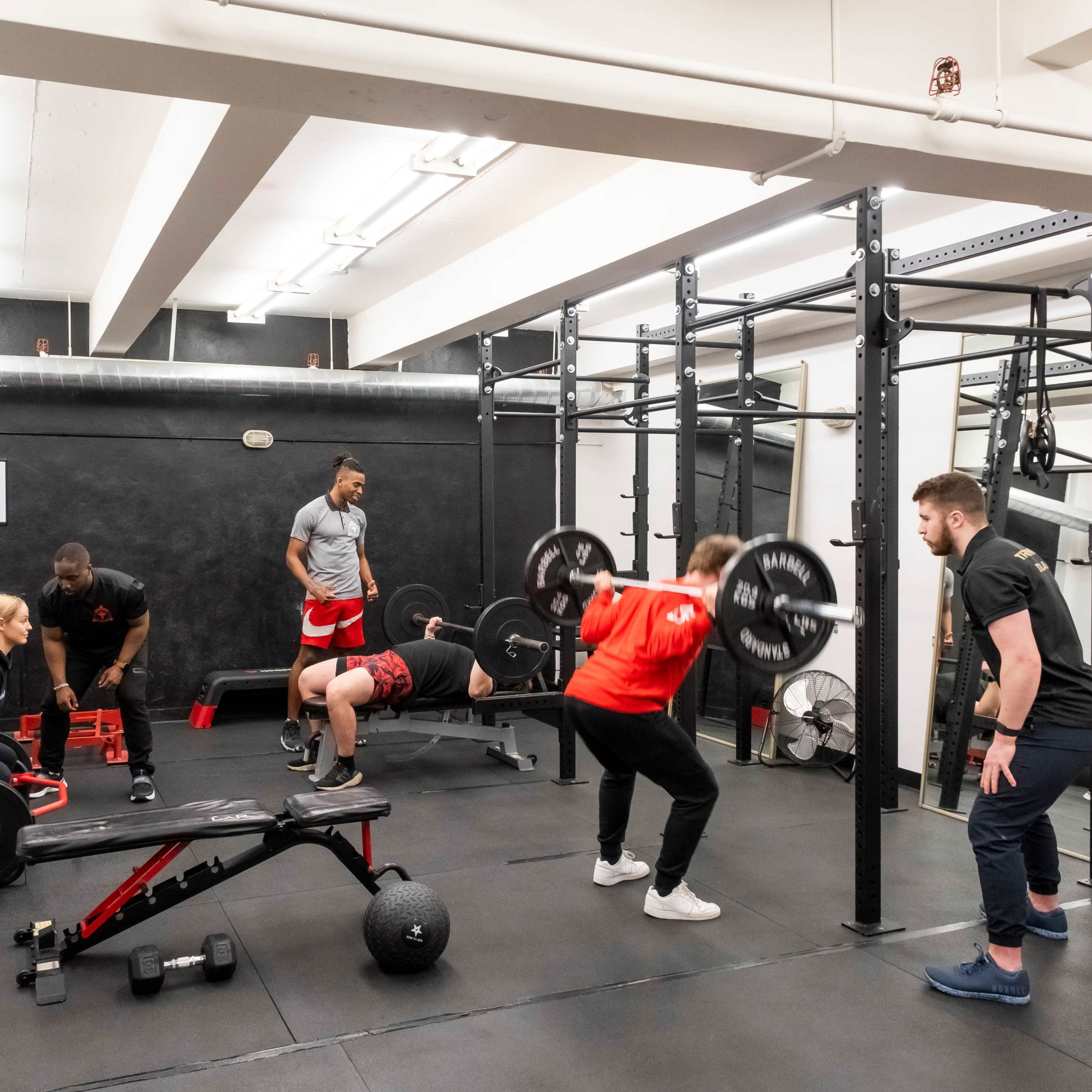 A group of individuals is engaged in various weightlifting exercises in a gym setting.