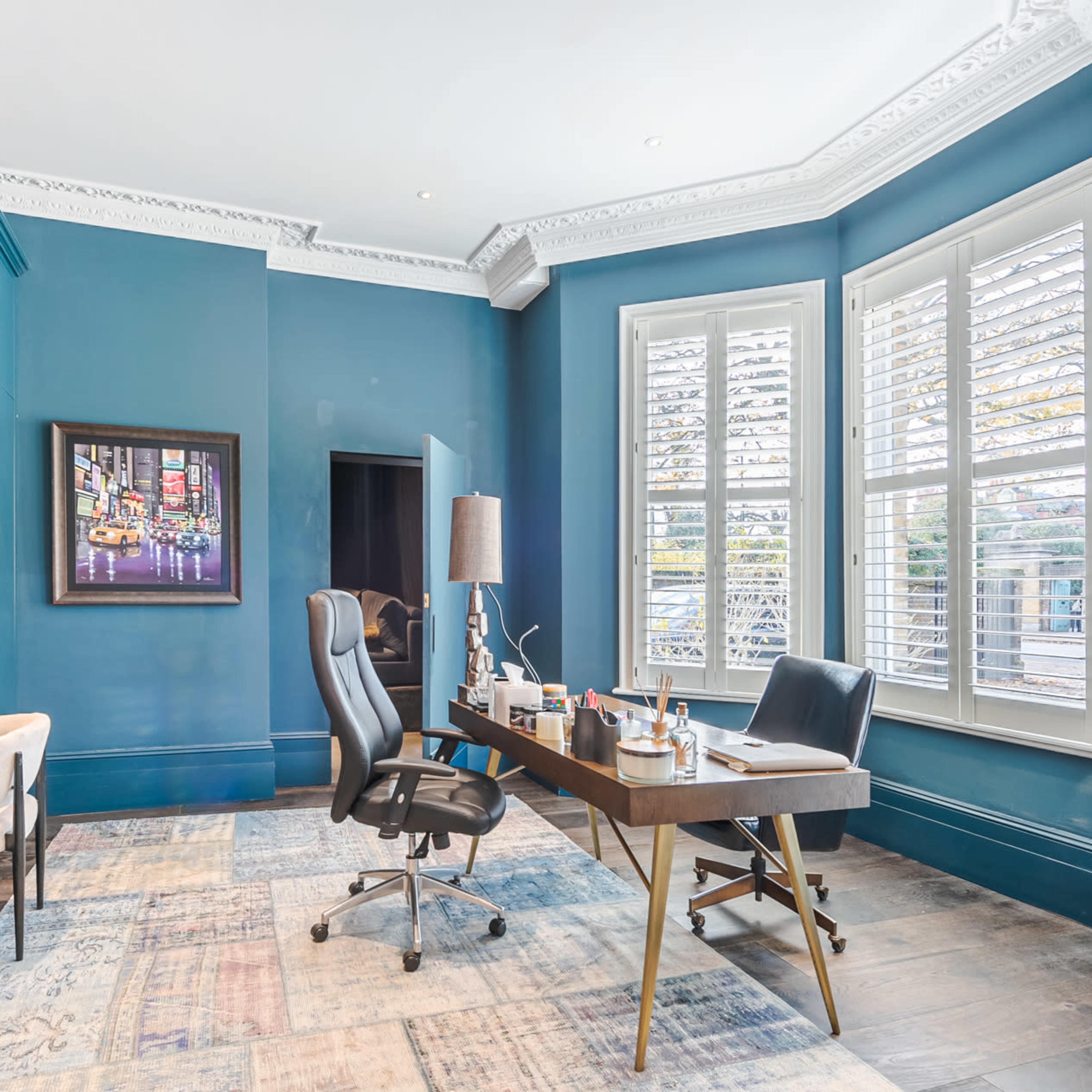 A modern office interior featuring a blue-painted wall, a wooden desk with office supplies, two chairs, and a bookshelf filled with books.