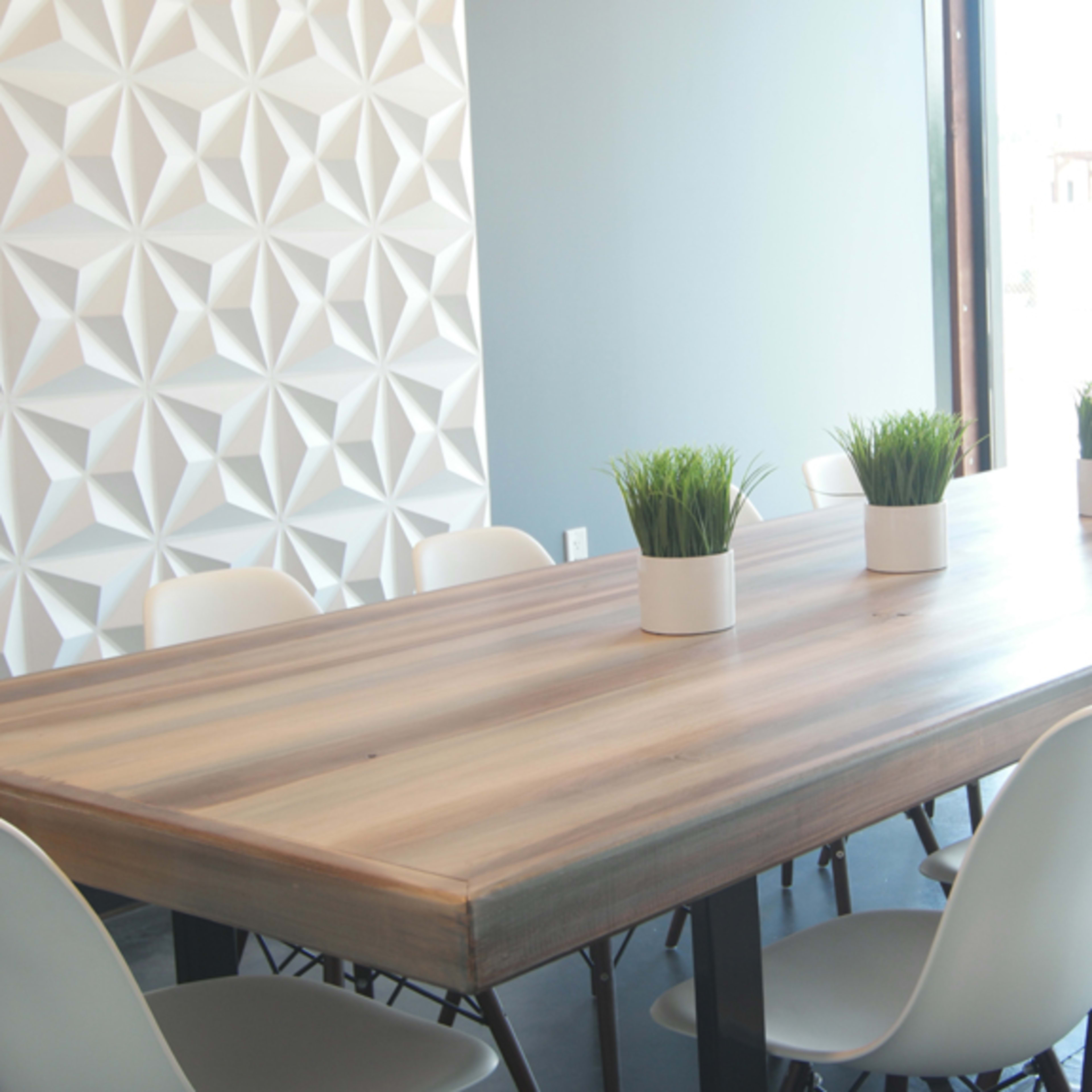 A long wooden conference table with potted plants sits in a bright room with a textured white wall and modern chairs.