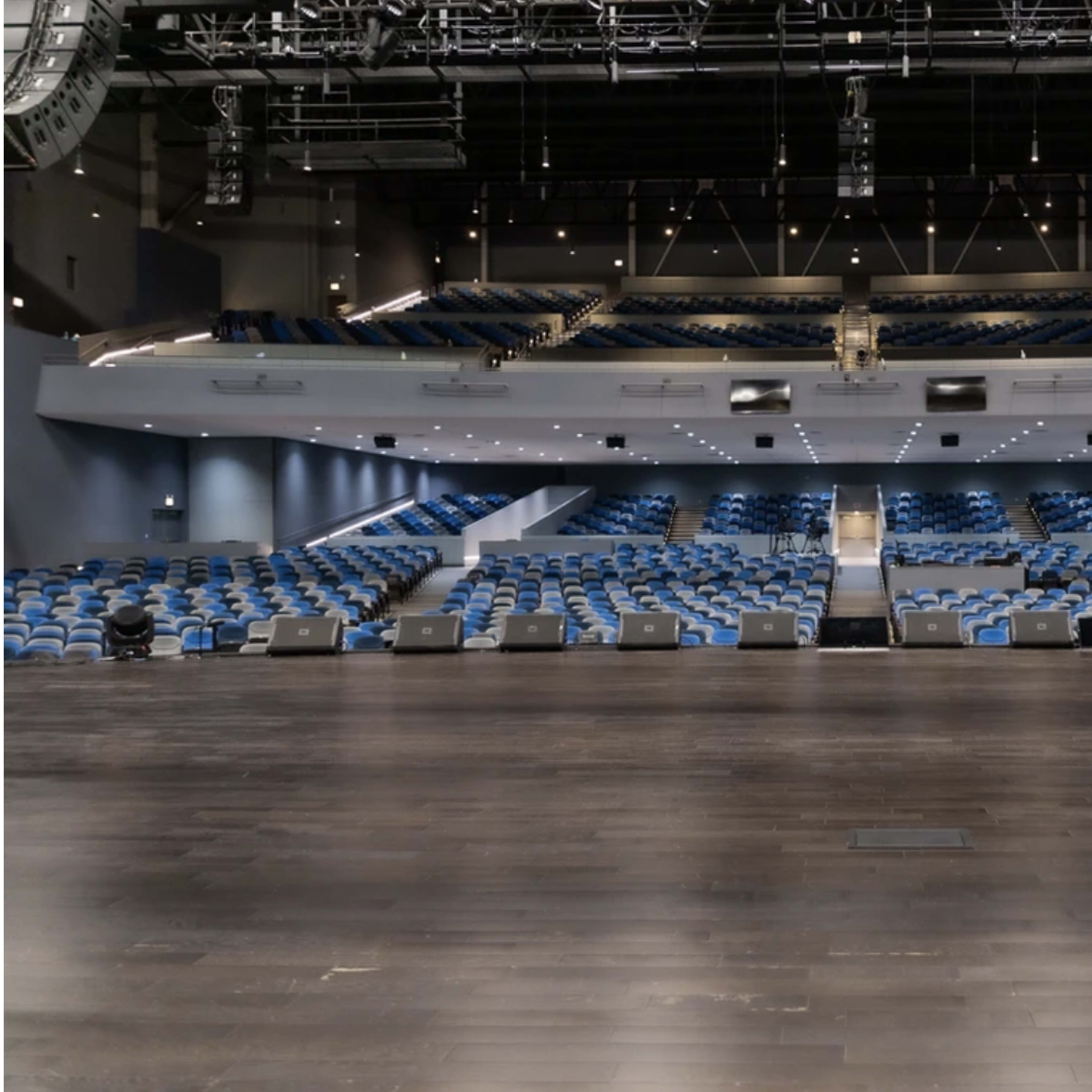 An empty auditorium with a wooden stage, blue seats arranged in tiered rows, and overhead stage lighting.