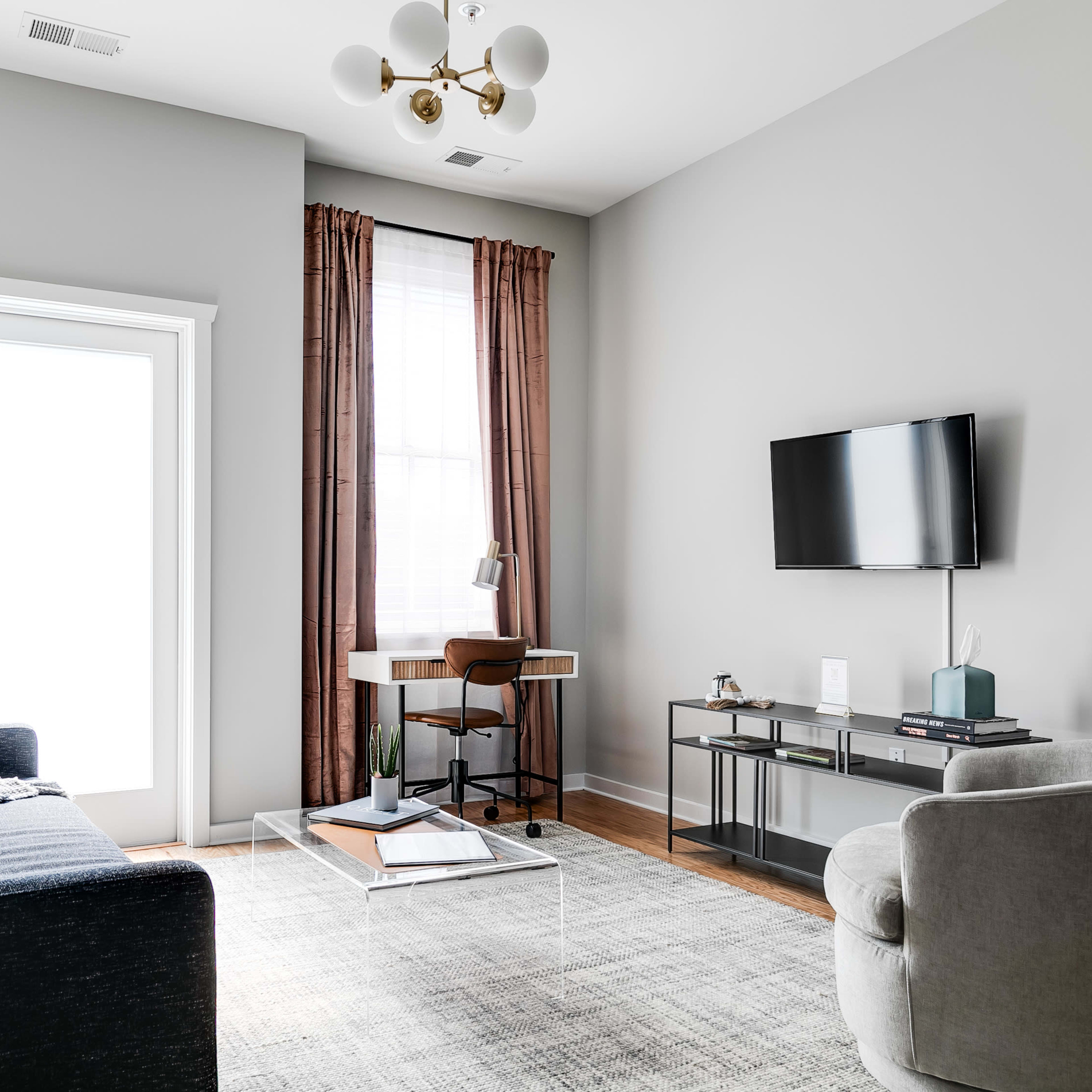 A modern living room with a blue sofa, a round chair, a glass coffee table, a desk with a chair, and a wall-mounted TV.