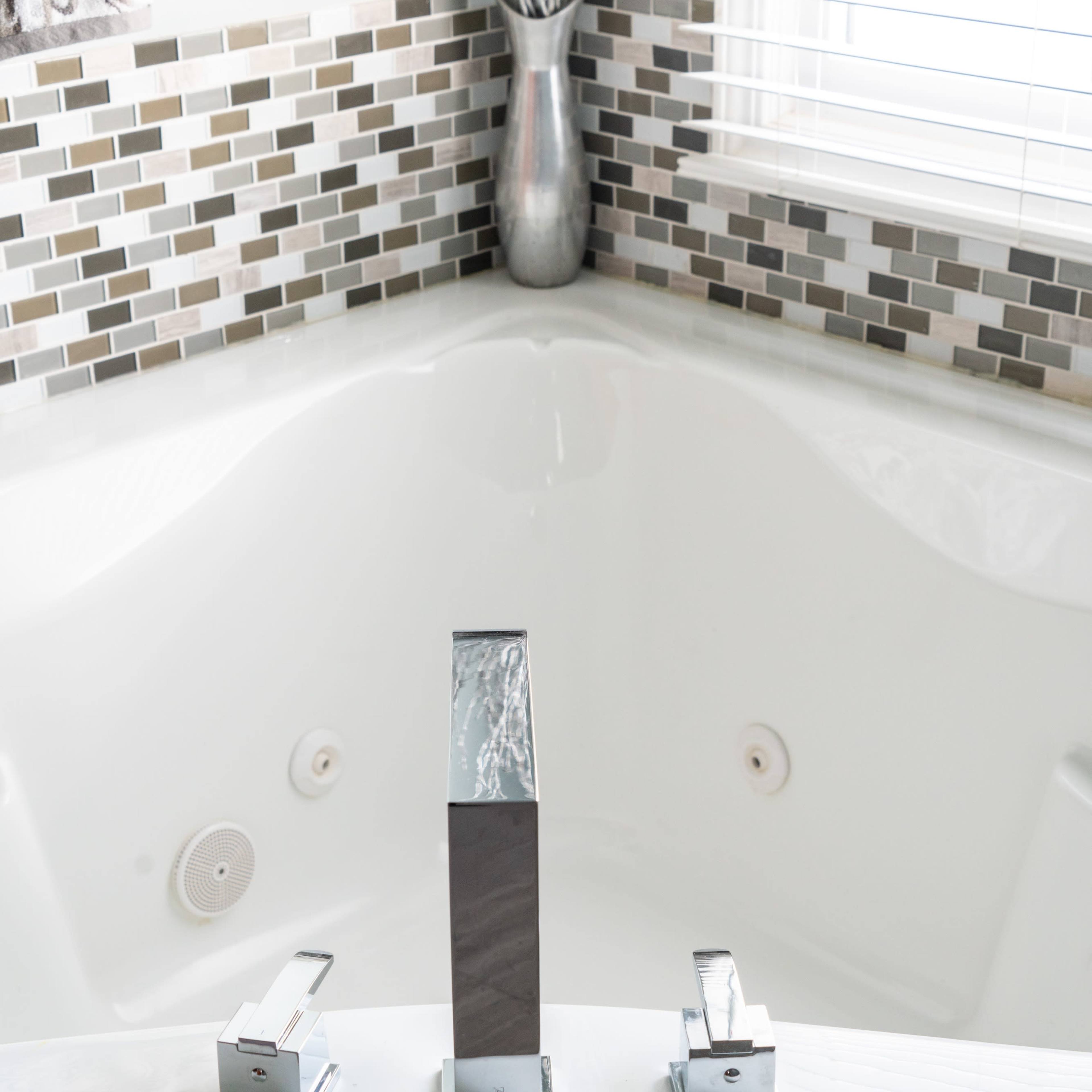 A modern bathtub with three chrome faucets and a mosaic tile backsplash.