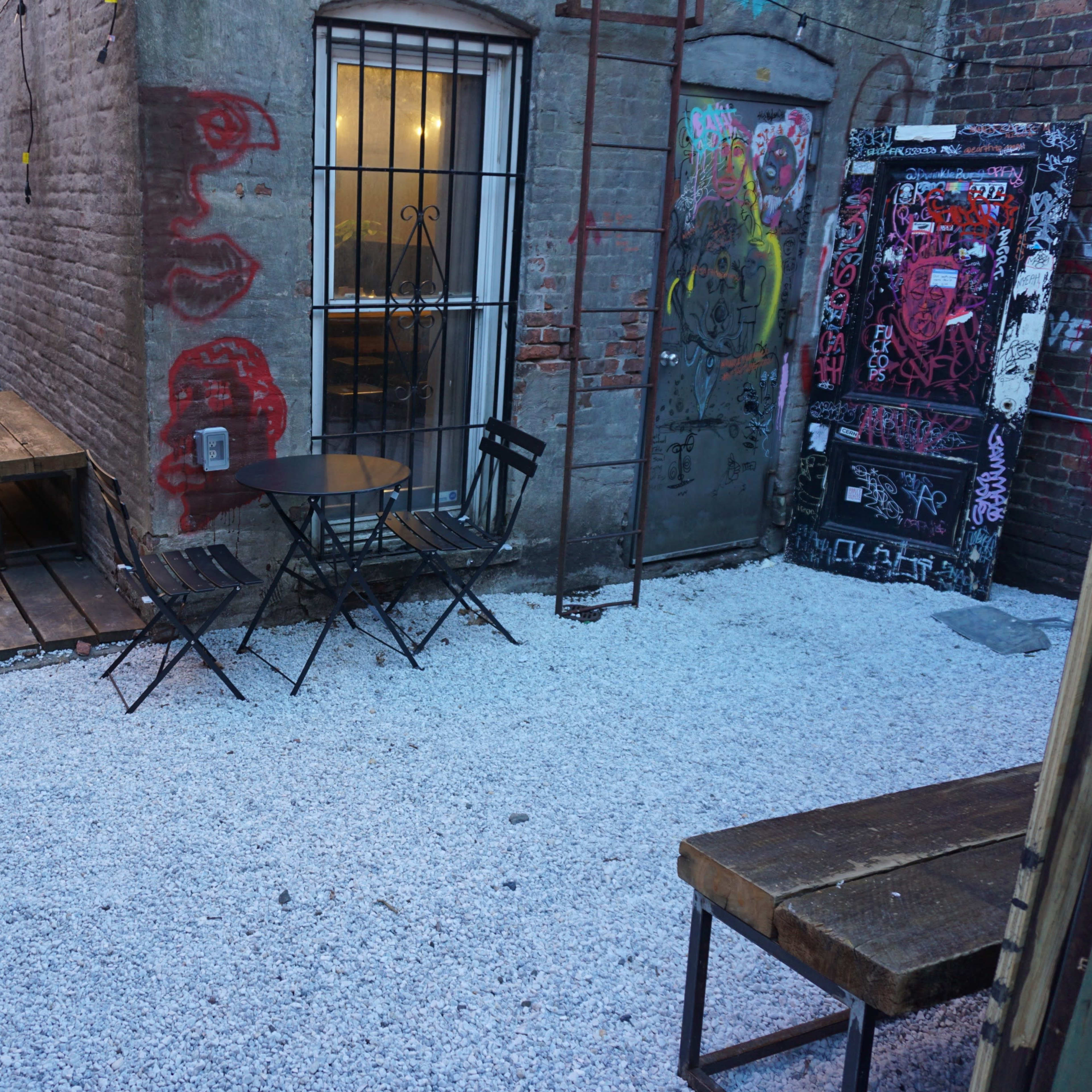 An alleyway with wooden and gravel surfaces, featuring metal tables and chairs surrounded by brick walls adorned with graffiti.