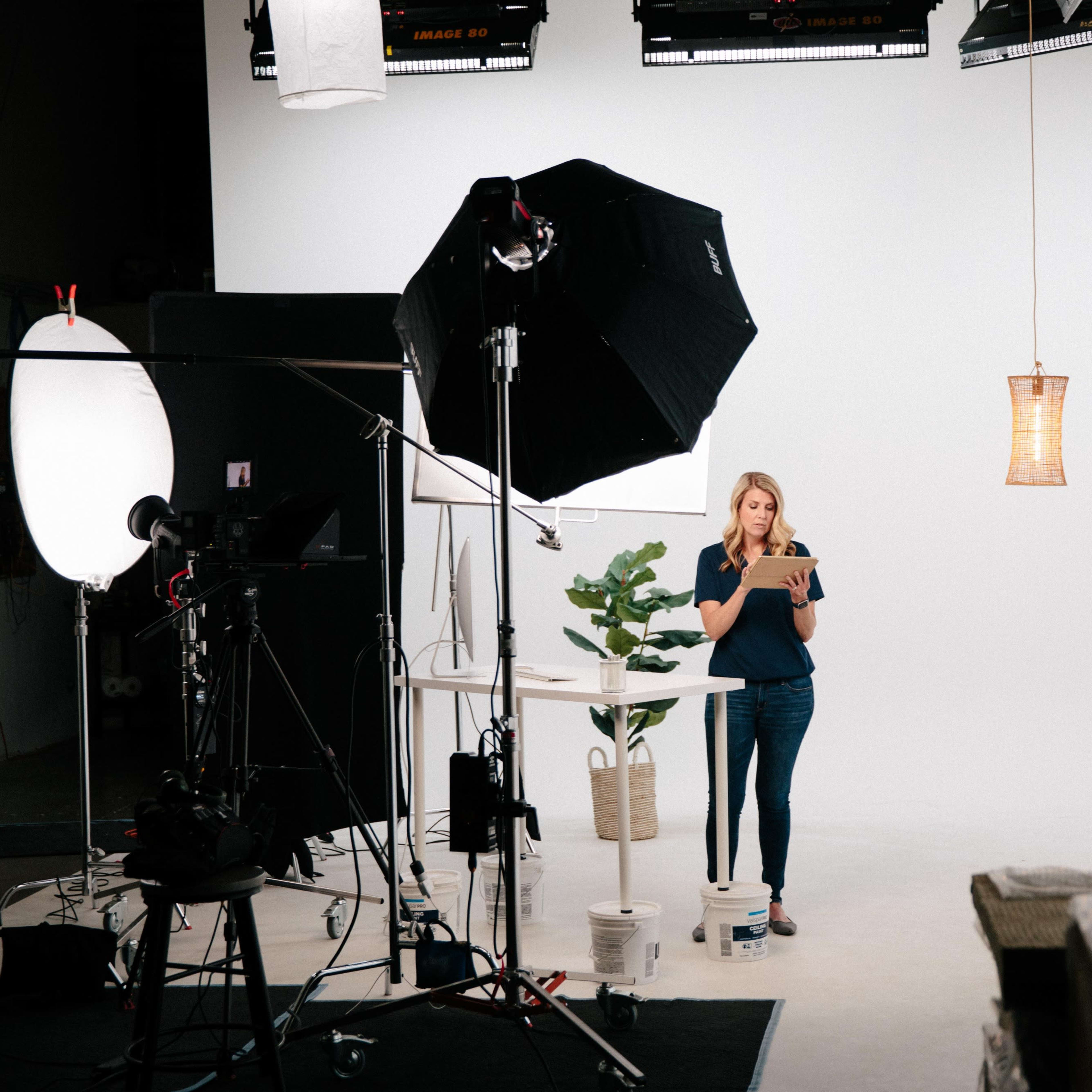 A woman stands on a set with various studio lights and camera equipment, holding a tablet while facing a blank white background.