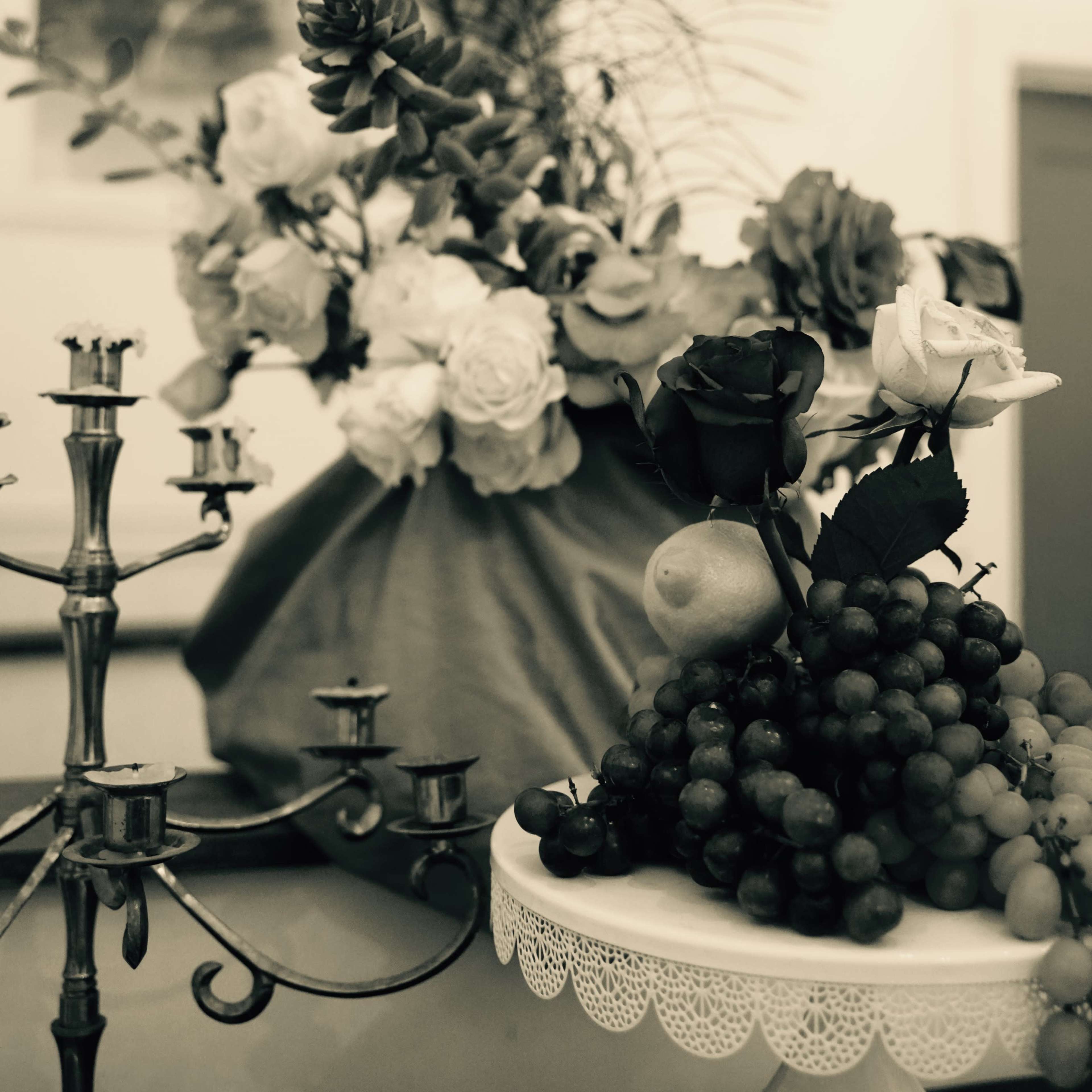 A table arrangement displays a candelabrum next to a fruit platter adorned with grapes and a pear, alongside a bouquet of flowers and peacock feathers.