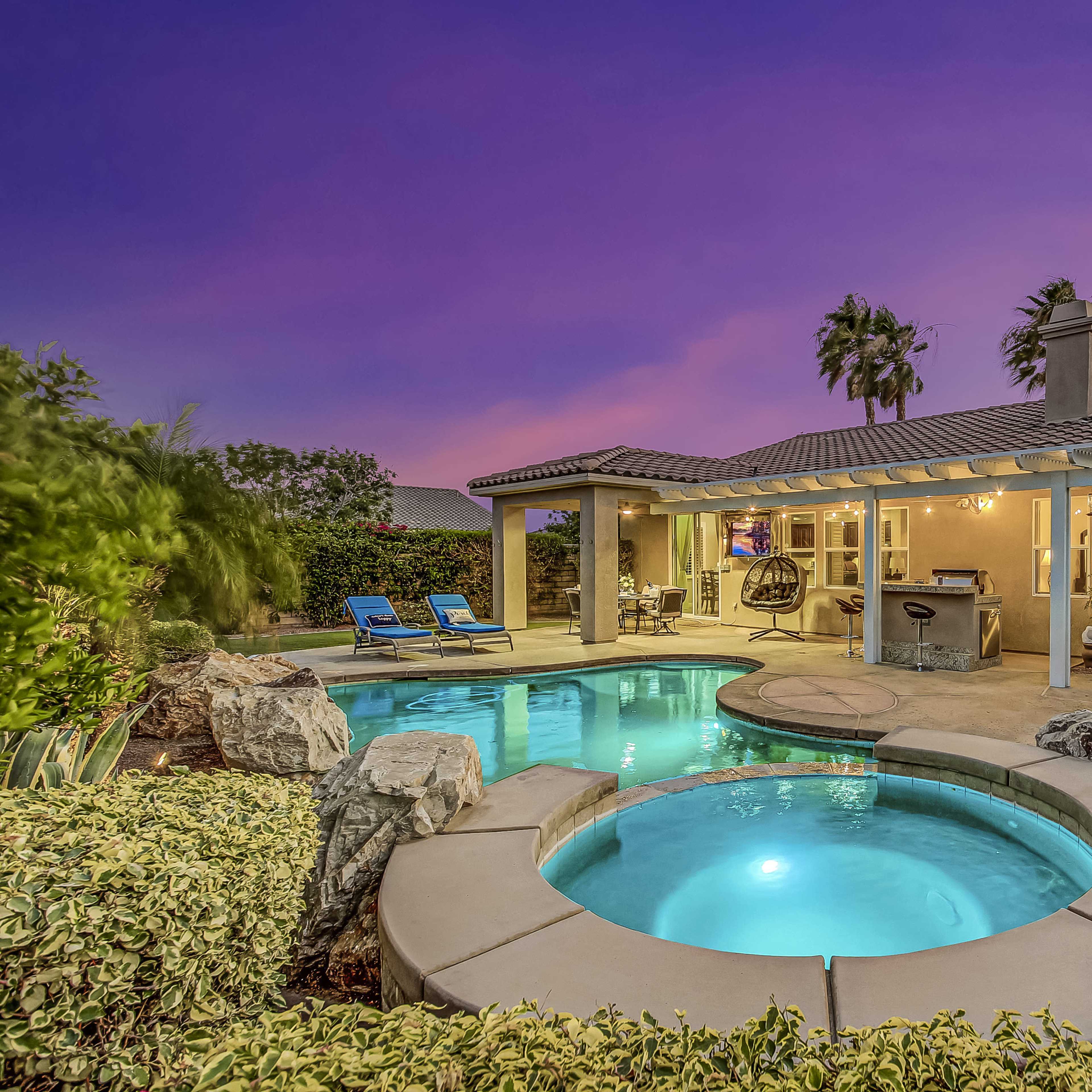 A backyard features a pool and a spa surrounded by palm trees and outdoor seating, with a home in the background under a twilight sky.
