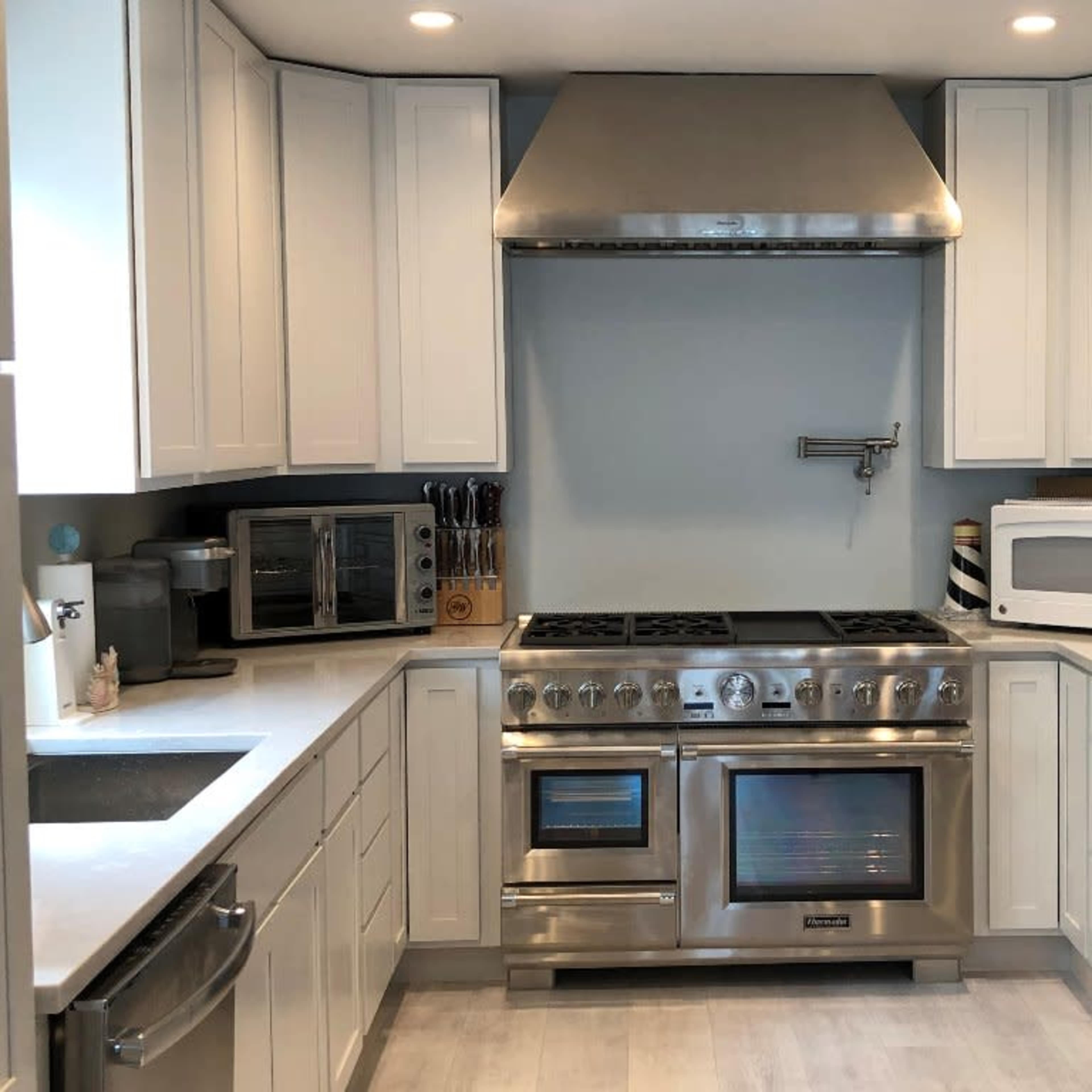 A modern kitchen with white cabinetry, a stainless steel range oven, a microwave, and various kitchen appliances neatly arranged on the countertops.