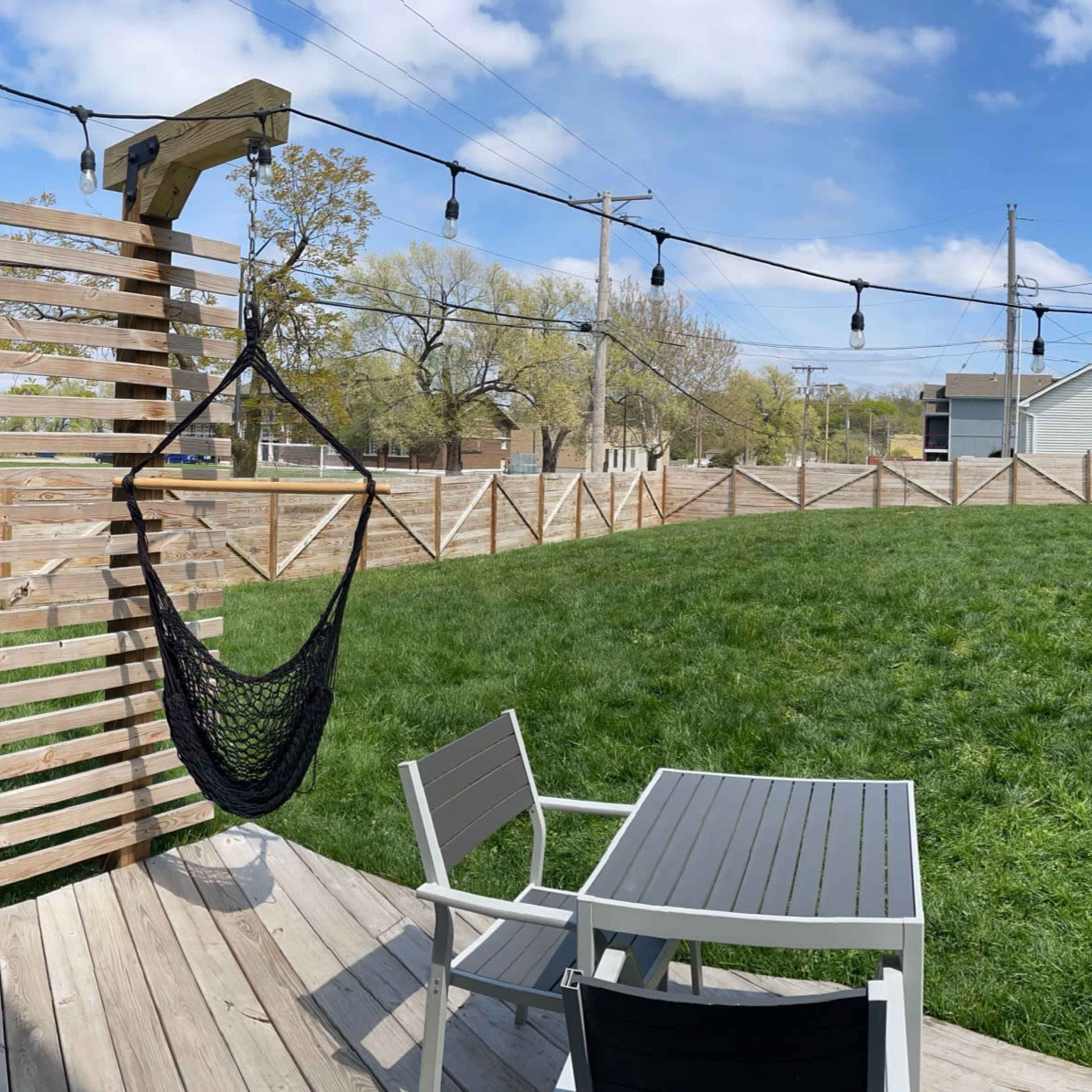 A backyard scene features a manicured lawn, a wooden deck with seating, a grill, and hanging string lights.