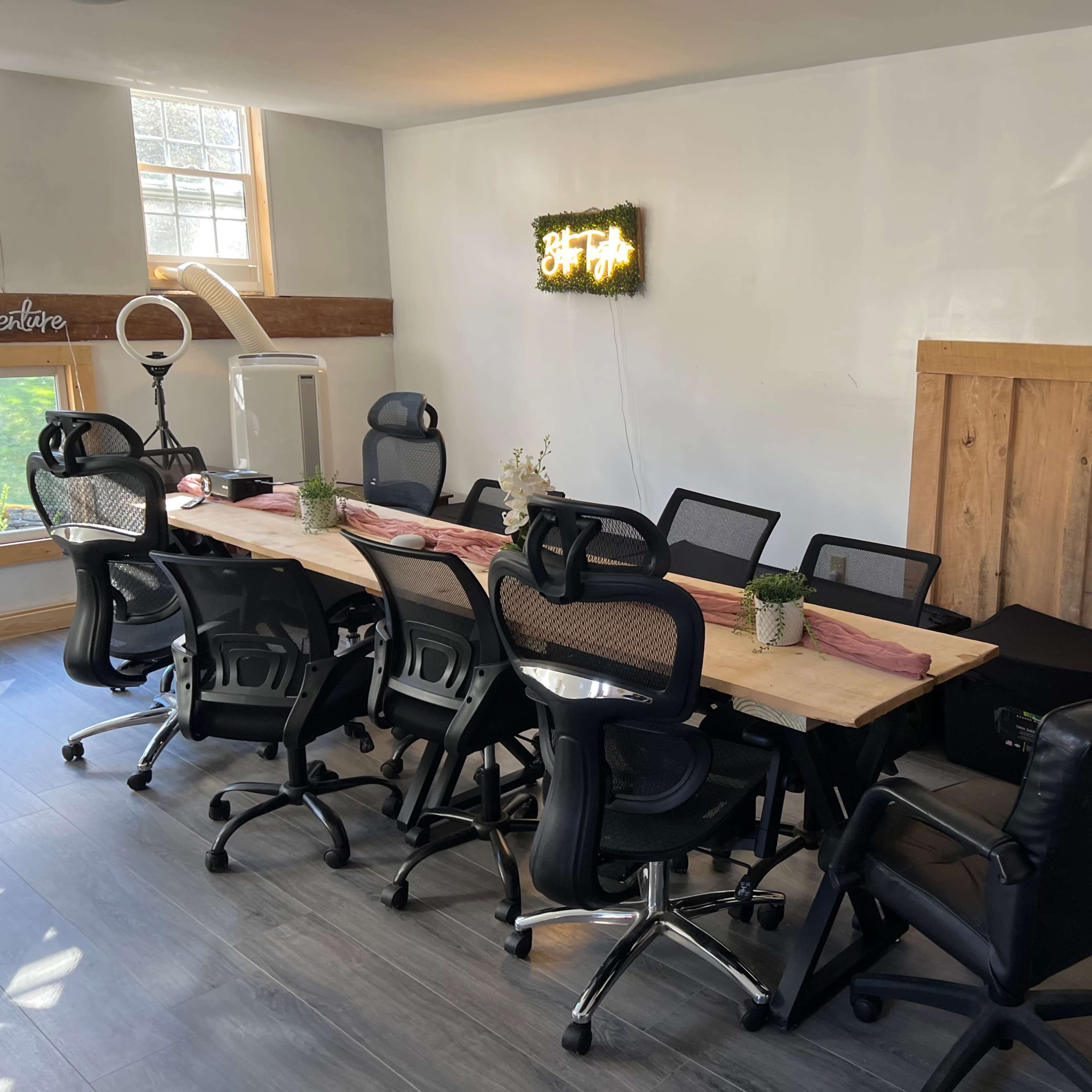 A modern conference room with a long wooden table surrounded by black office chairs, a mini fridge, and a neon sign on the wall.