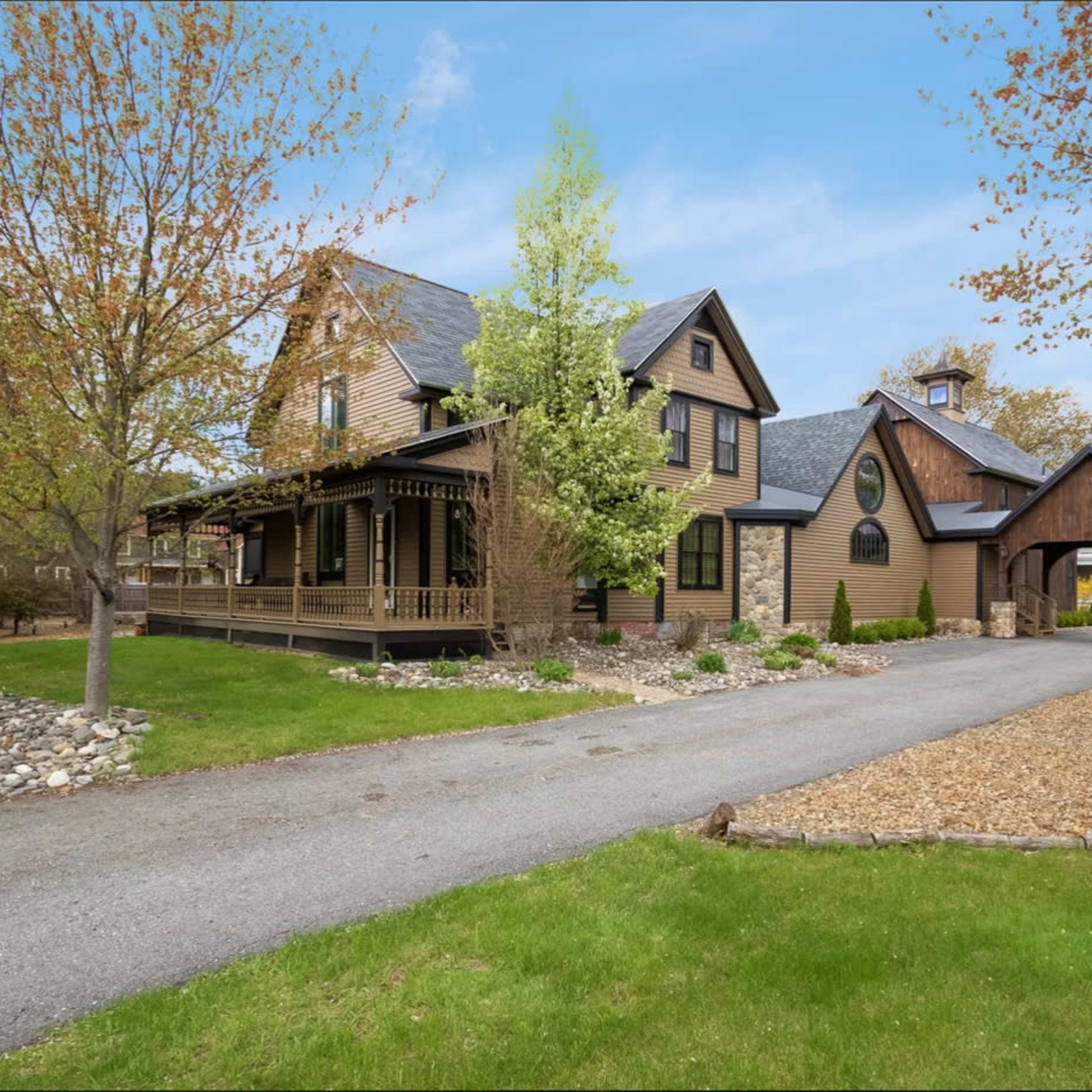 A two-story house with a mix of wood and stone exterior, situated beside a gravel driveway and landscaped yard with trees and shrubs.