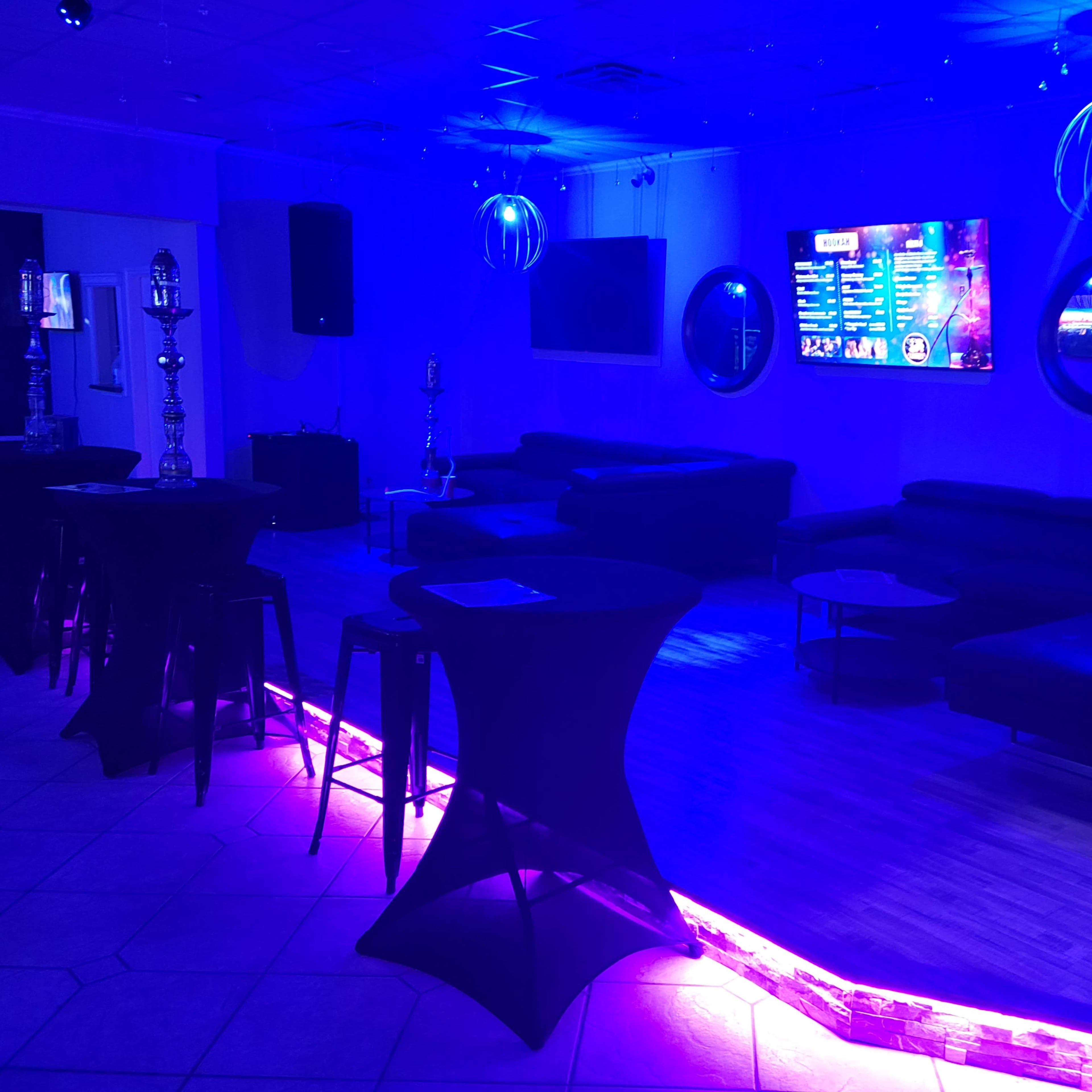 A lounge area with low seating, illuminated by blue and purple lights, and equipped with screens displaying information or entertainment.