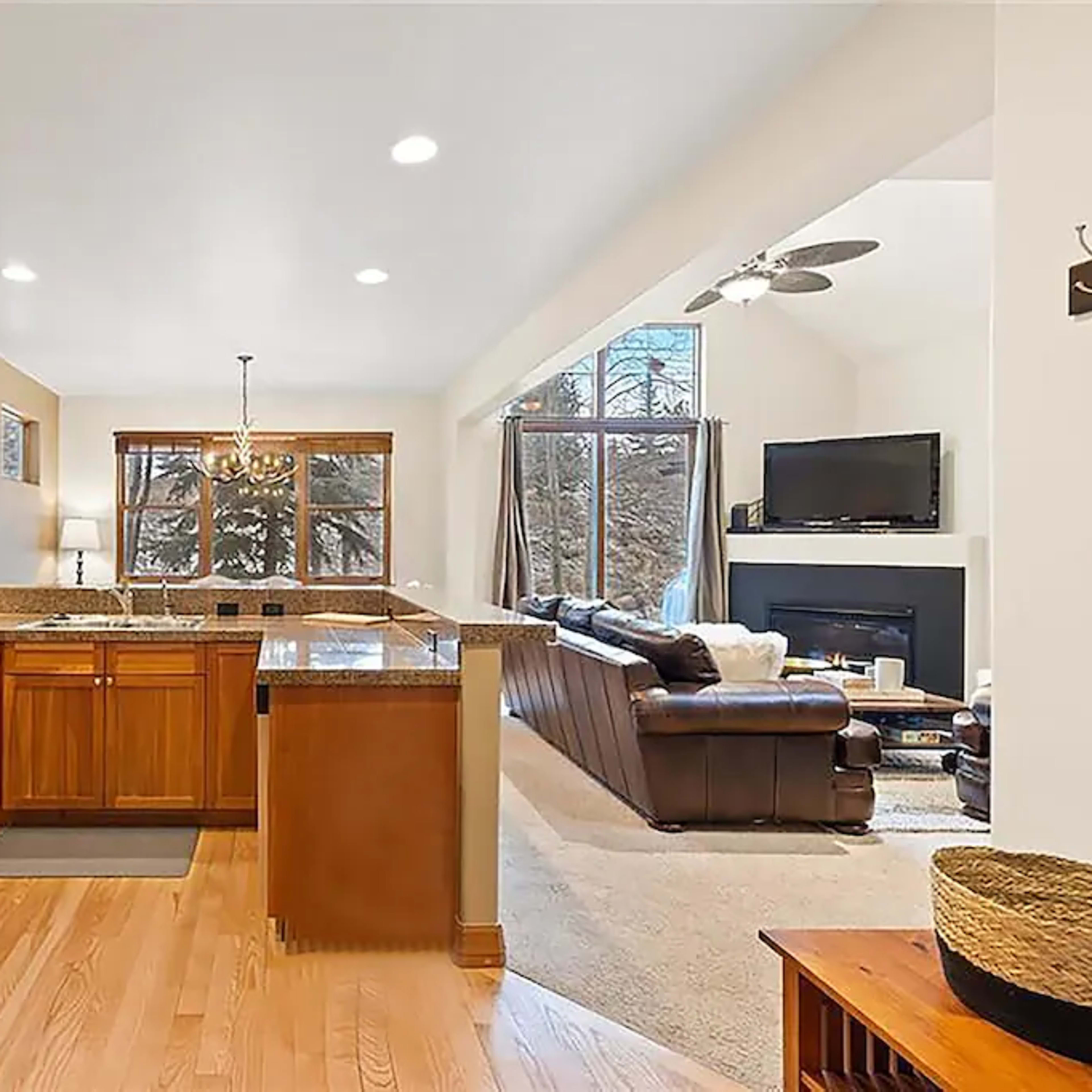 A modern kitchen features wooden cabinets, granite countertops, and stainless steel appliances, connected to a living area with large windows and a fireplace.