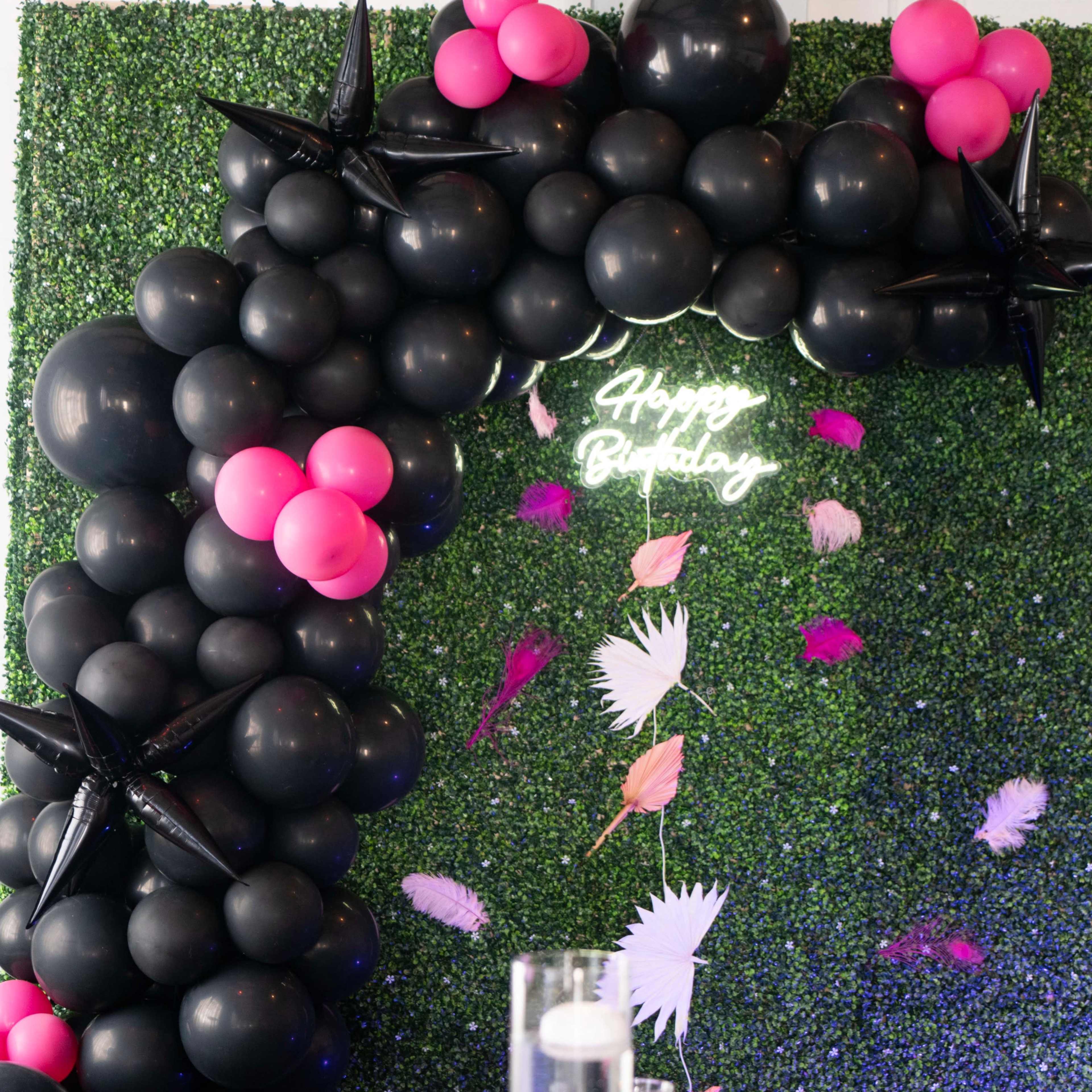 A decorative backdrop featuring black and pink balloons arranged in an arch with a neon "Happy Birthday" sign against a green foliage wall.