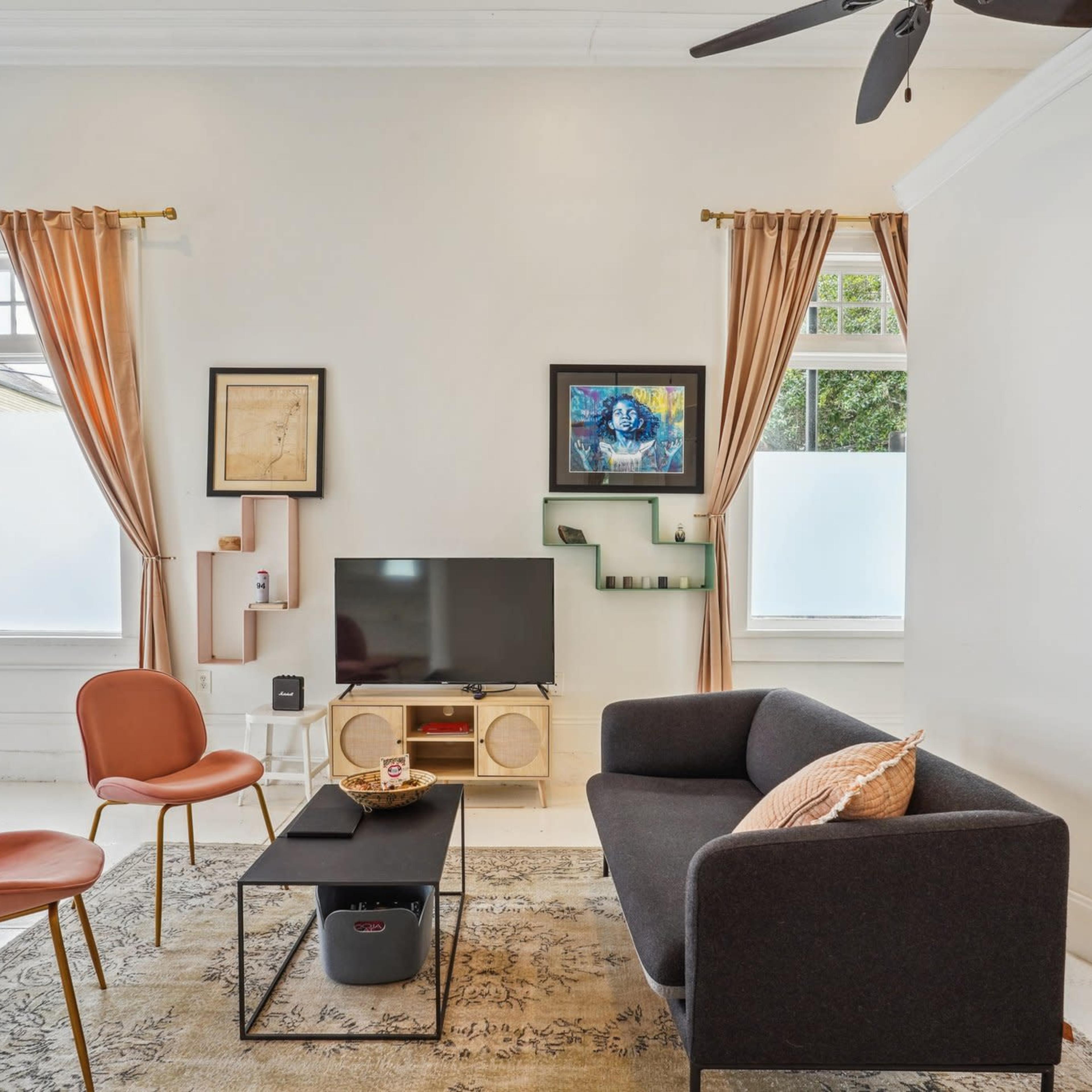 A living room features a dark couch, two pink chairs, a low coffee table, and a wall-mounted TV with artwork displayed nearby.