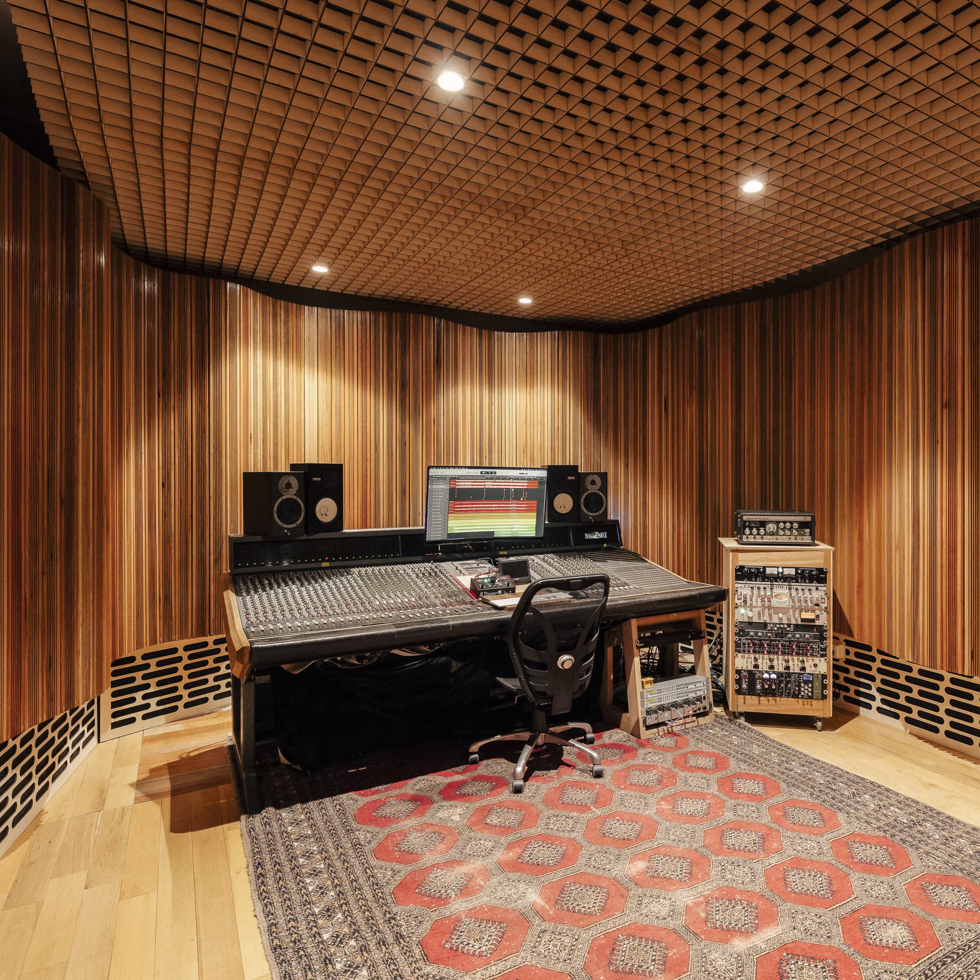 A music studio features a wooden-paneled interior with a central mixing desk, speakers, and electronic equipment.