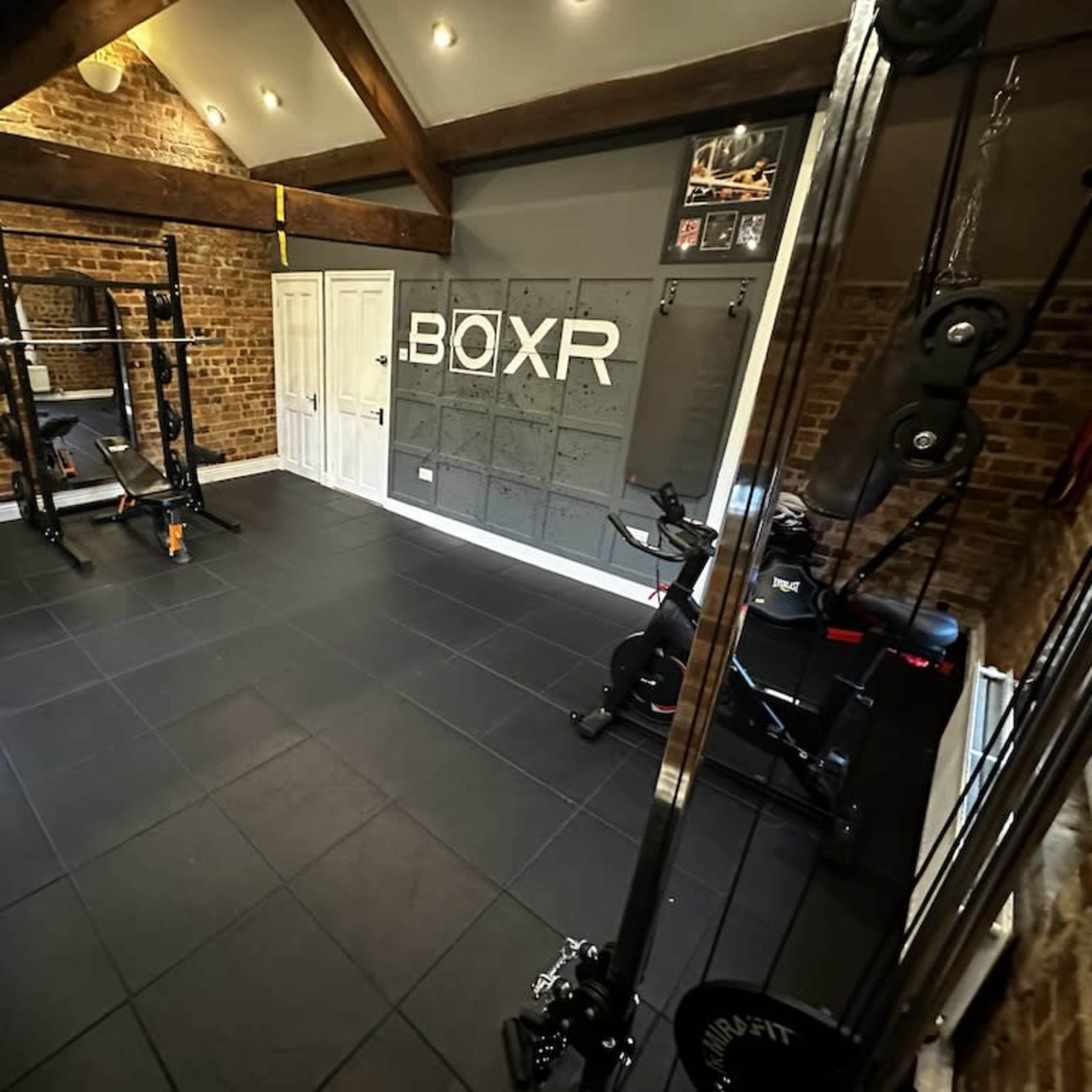 A well-equipped home gym featuring a squat rack, weight plates, a cable machine, and a stationary bike, all set against a backdrop of exposed brick walls and wooden beams.