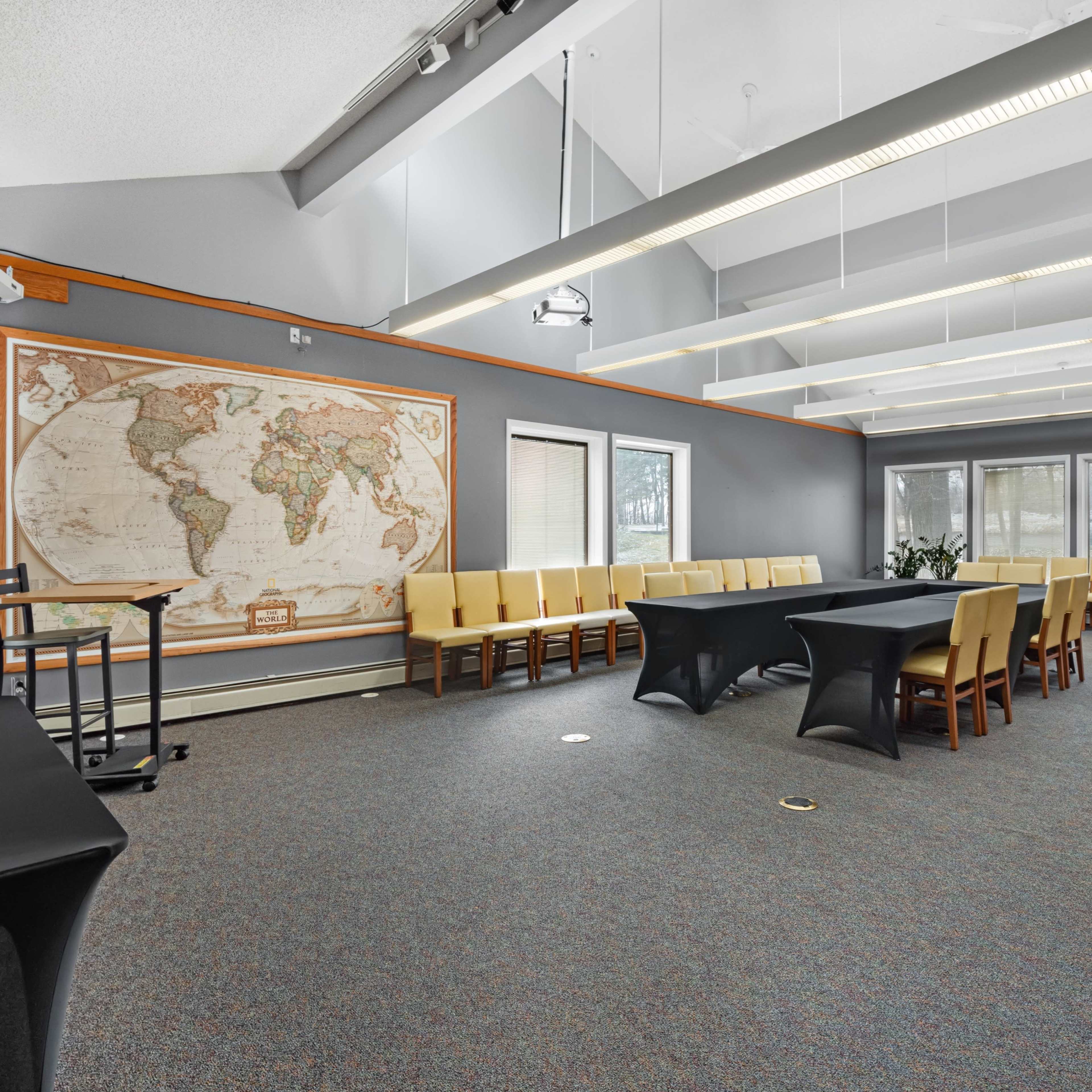 A well-lit conference room featuring a large world map on the wall, a central black table surrounded by chairs, and windows allowing natural light.