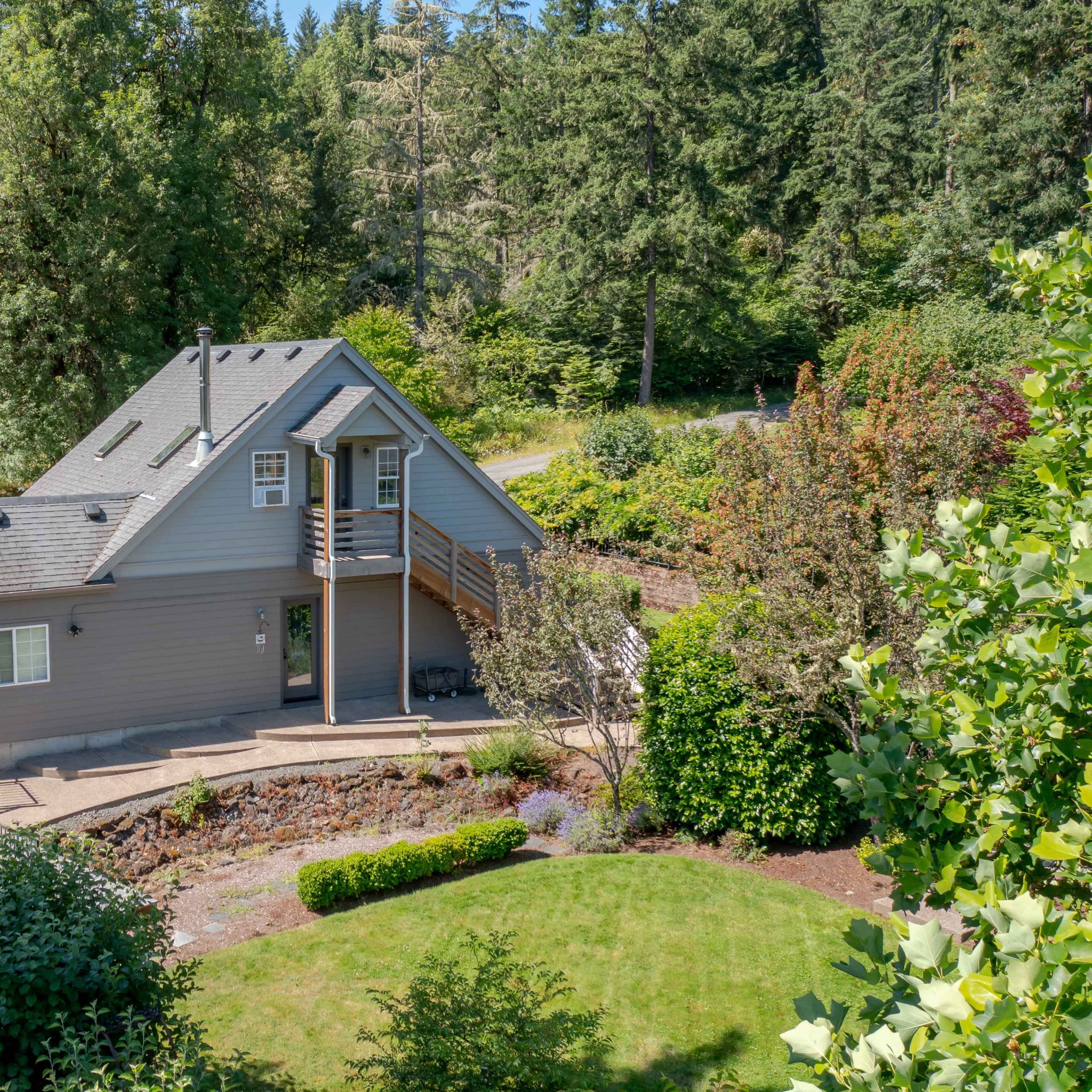 A two-story gray house surrounded by greenery and trees with a gravel driveway visible in the background.