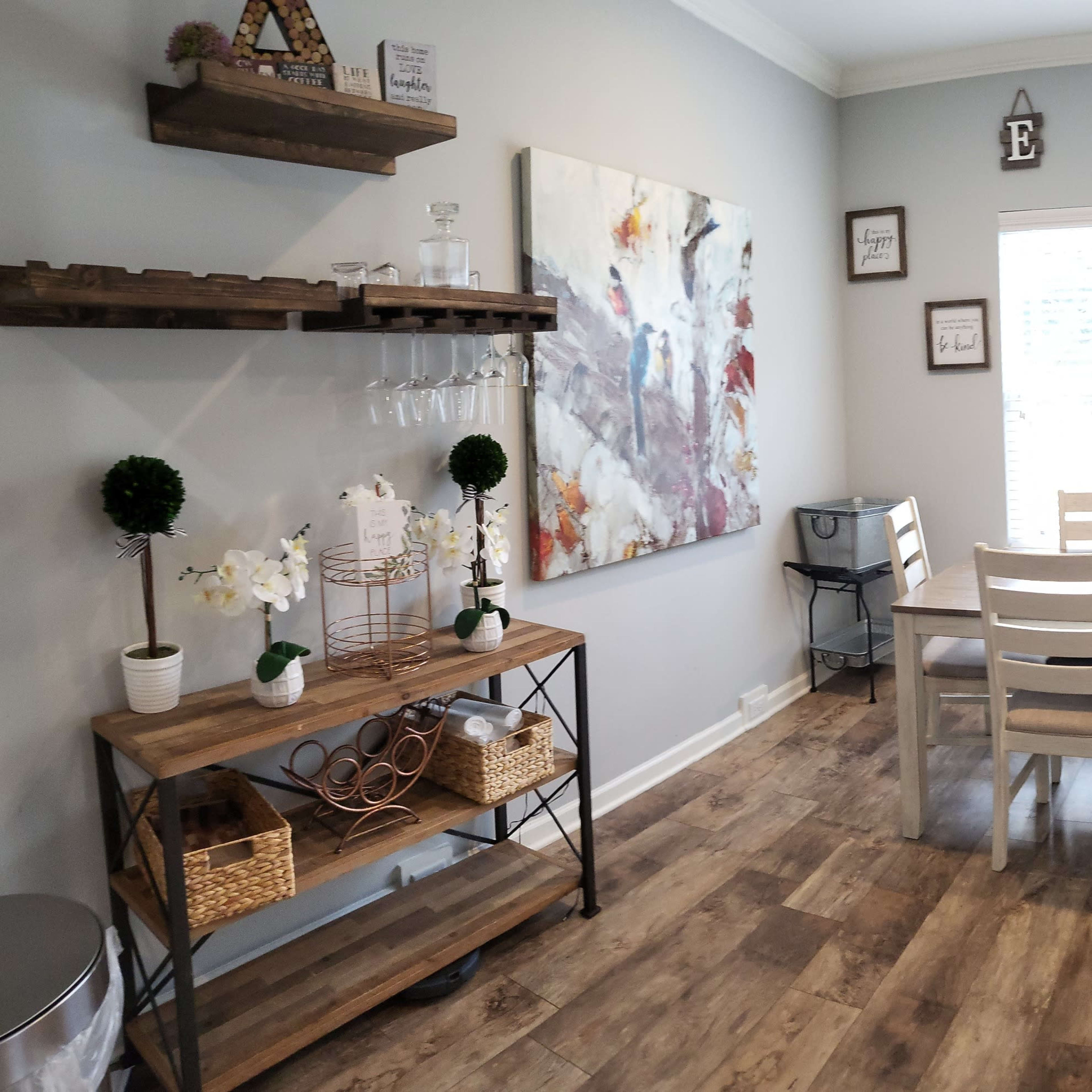 A dining area features a wooden table with white chairs, a shelf with decorative items, and a large window with blinds.