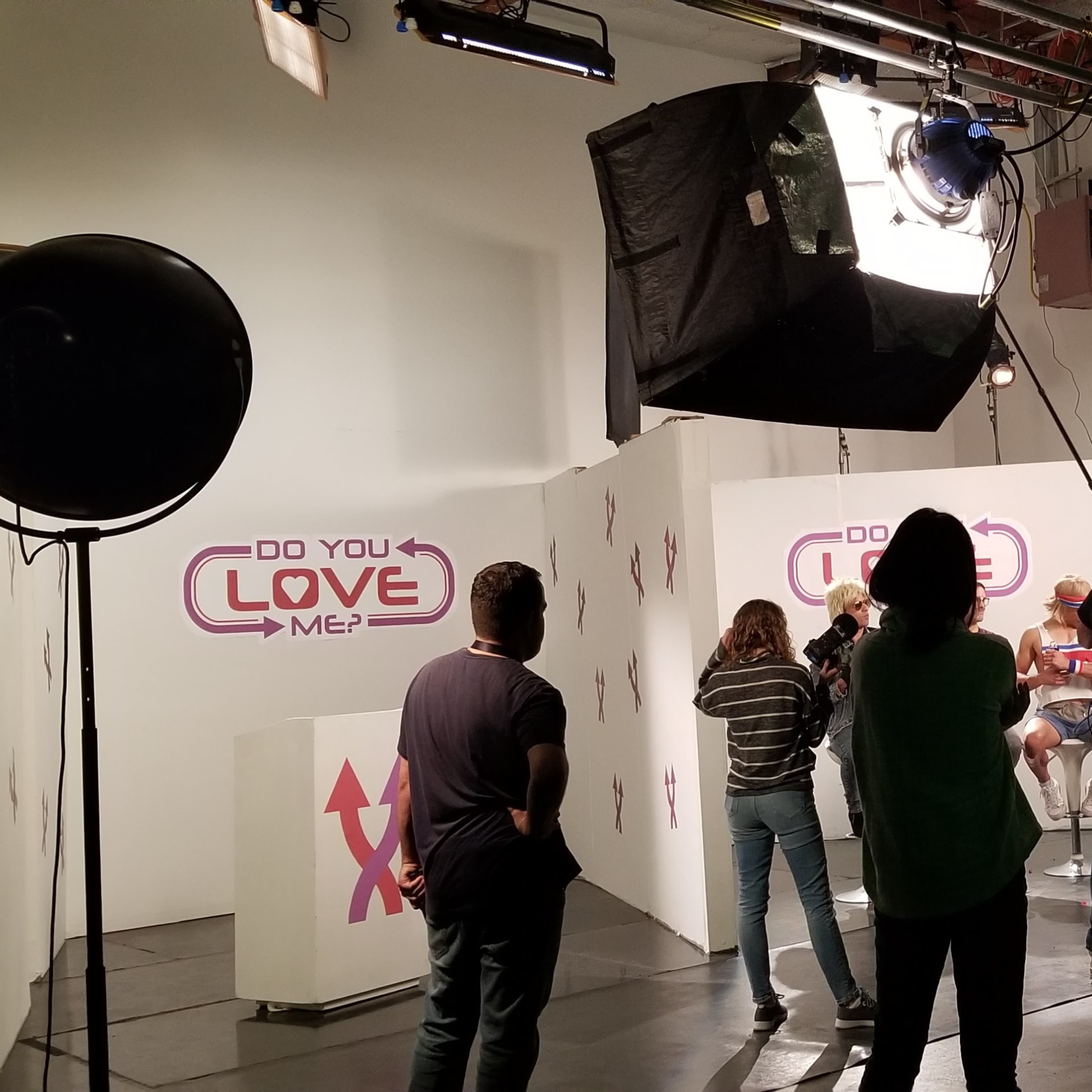 A television production crew prepares to film a segment in a studio with a backdrop that reads "DO YOU LOVE ME?"