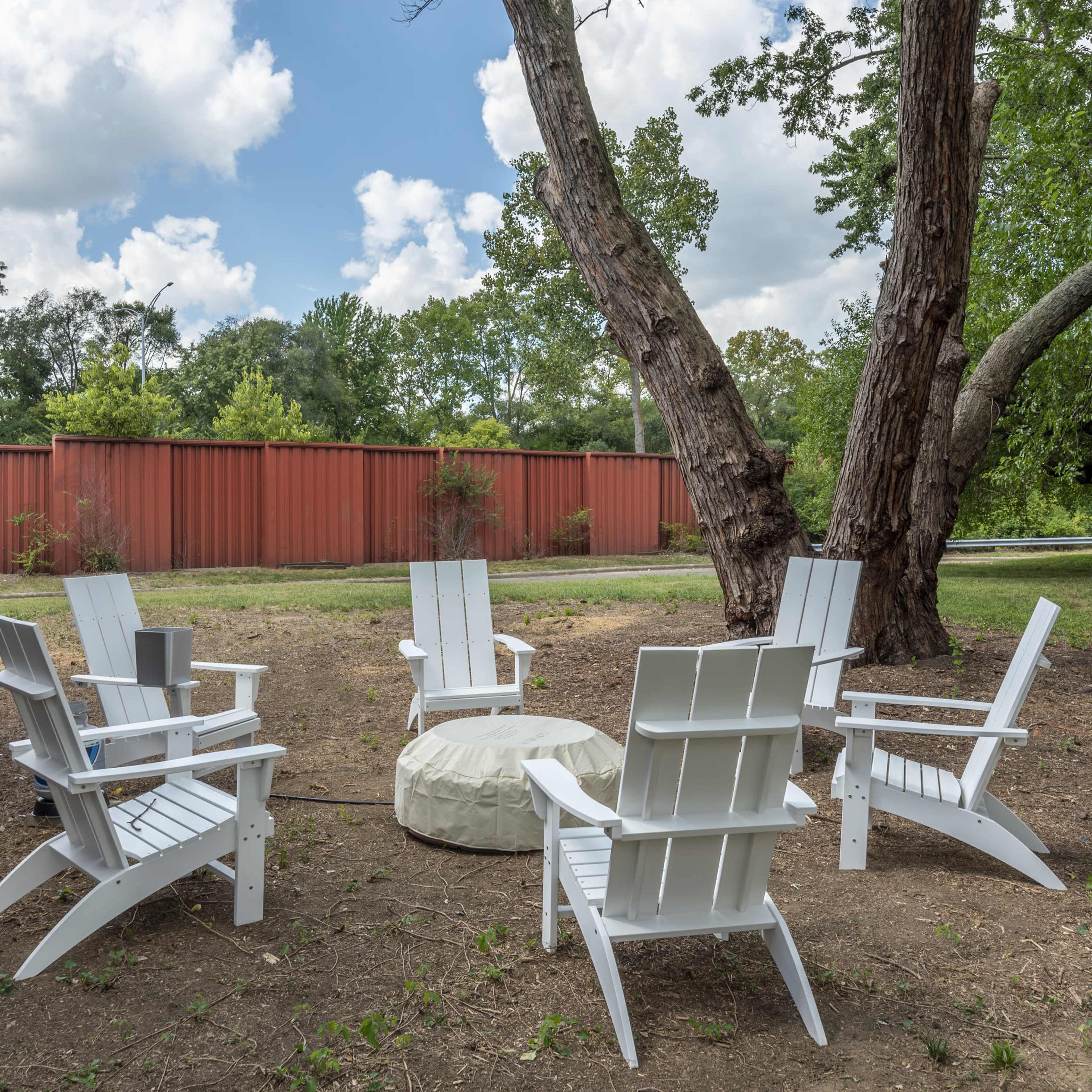 A group of five white Adirondack chairs surrounds a circular fire pit in a grassy area under trees.