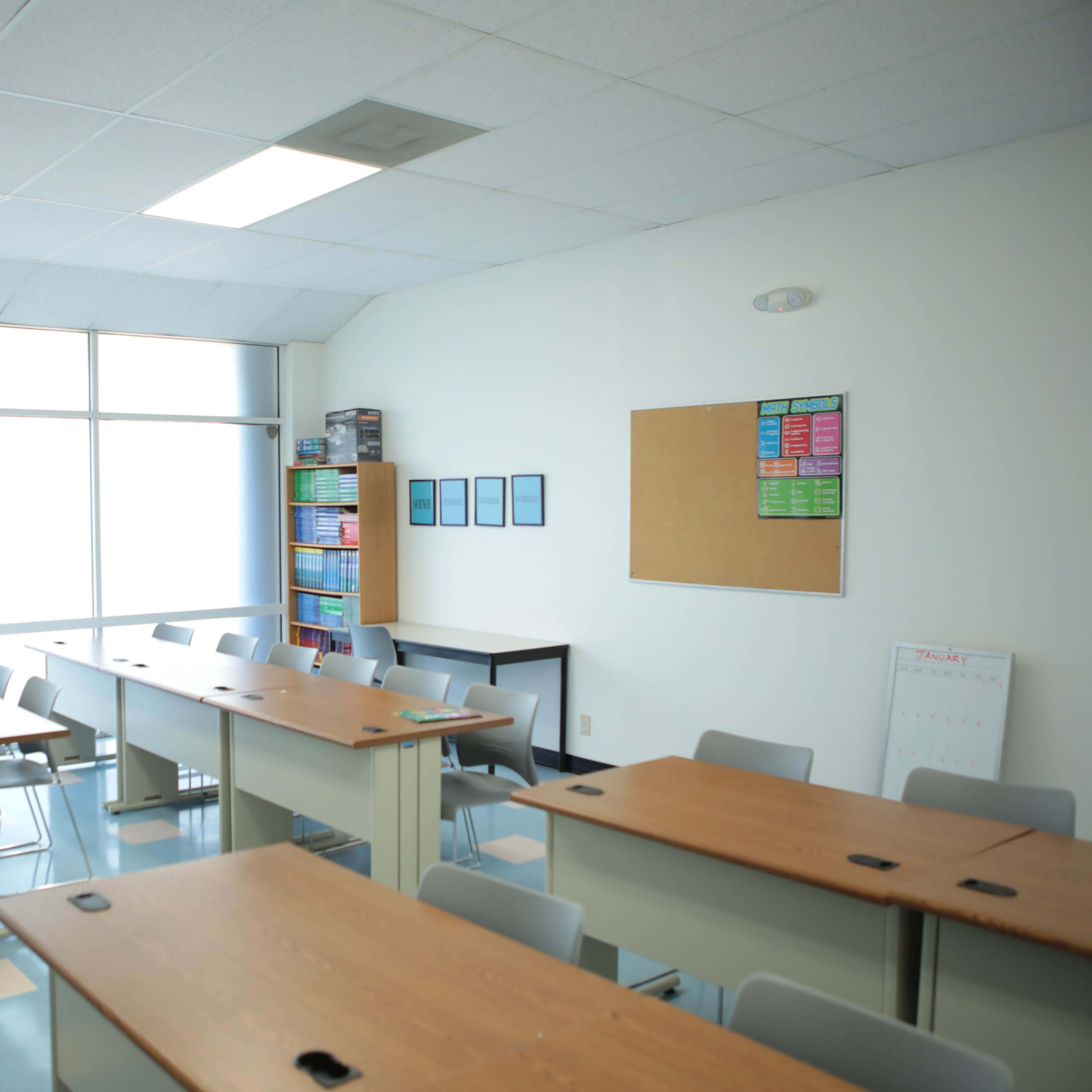 A classroom is arranged with several tables and chairs, featuring a whiteboard, a bulletin board, and shelves filled with books.