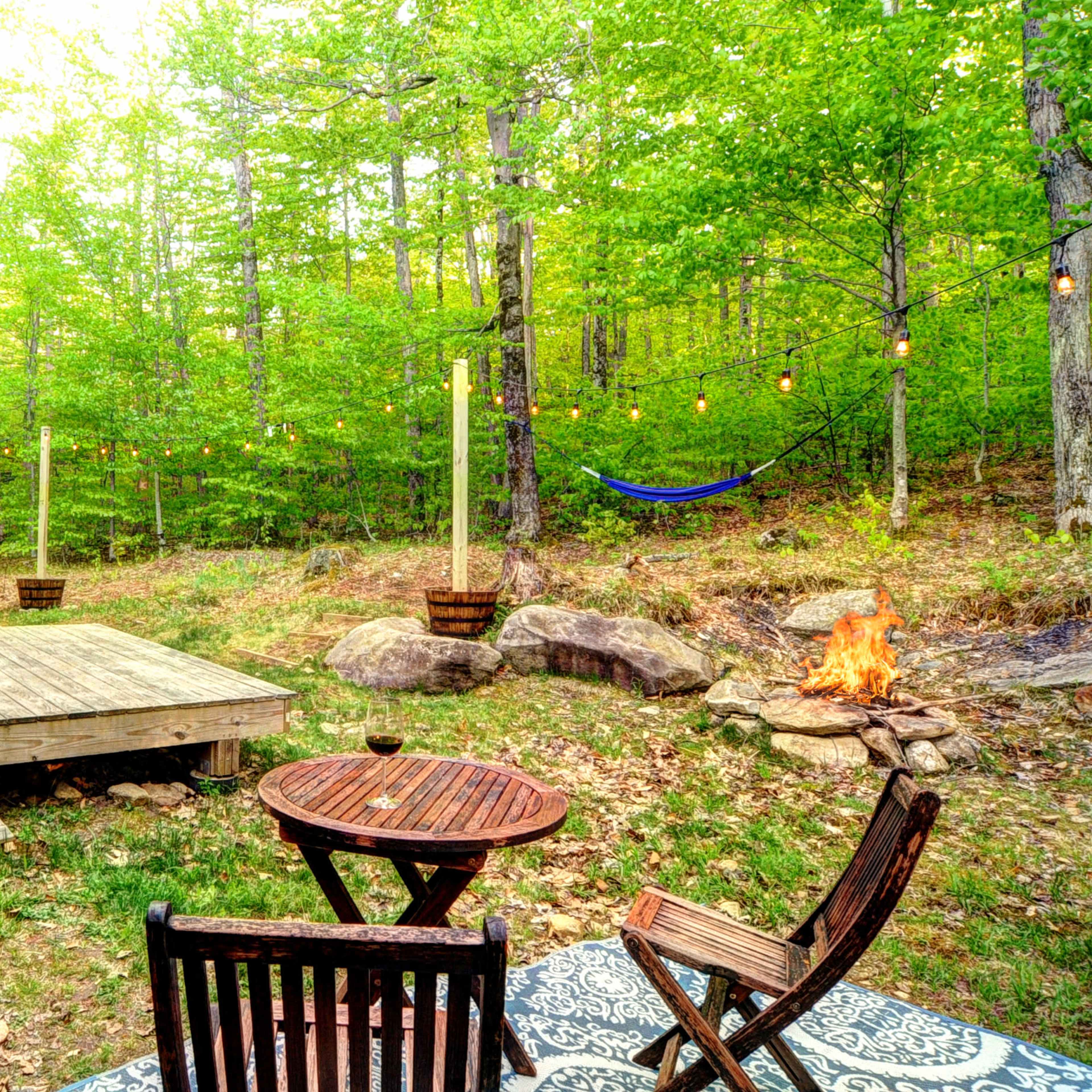 A wooden deck with a table and chairs is adjacent to a grill, overlooking a forested area featuring a hammock and a fire pit surrounded by rocks.