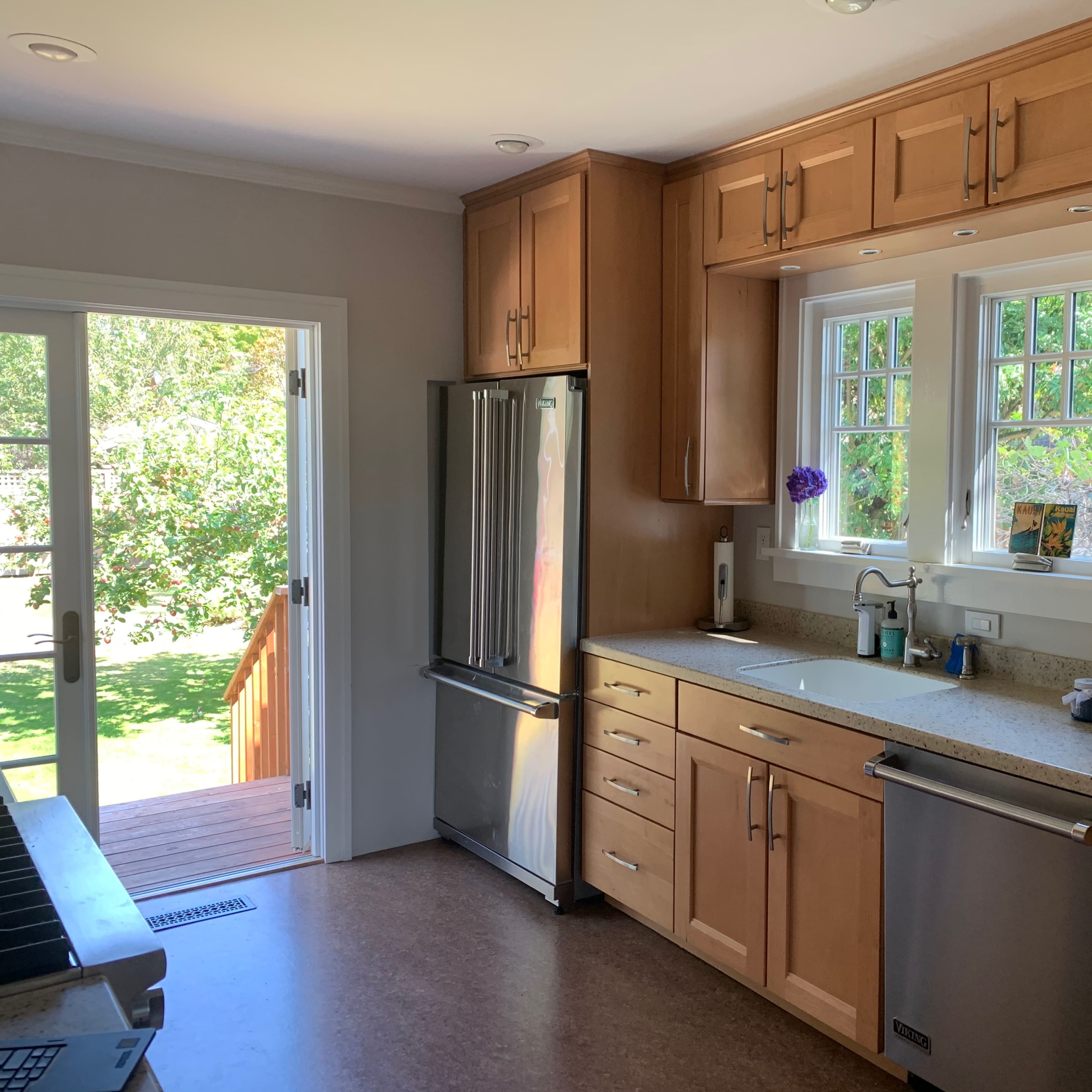 The kitchen features wooden cabinets, a stainless steel refrigerator, and a countertop with a sink and coffee maker, with a view of the outdoors through a doorway.