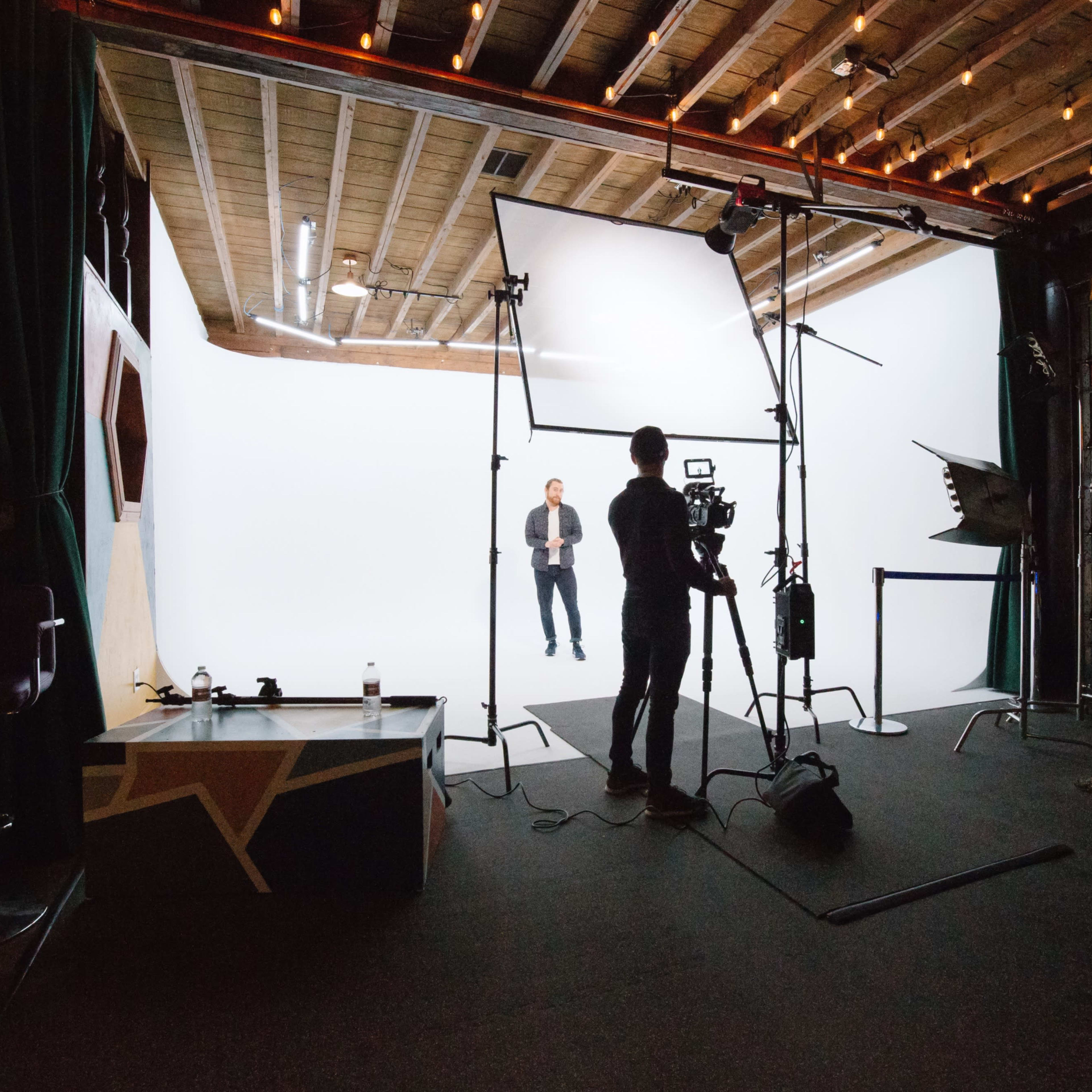 A person stands on a white backdrop inside a studio, while someone else sets up a camera and lighting equipment.