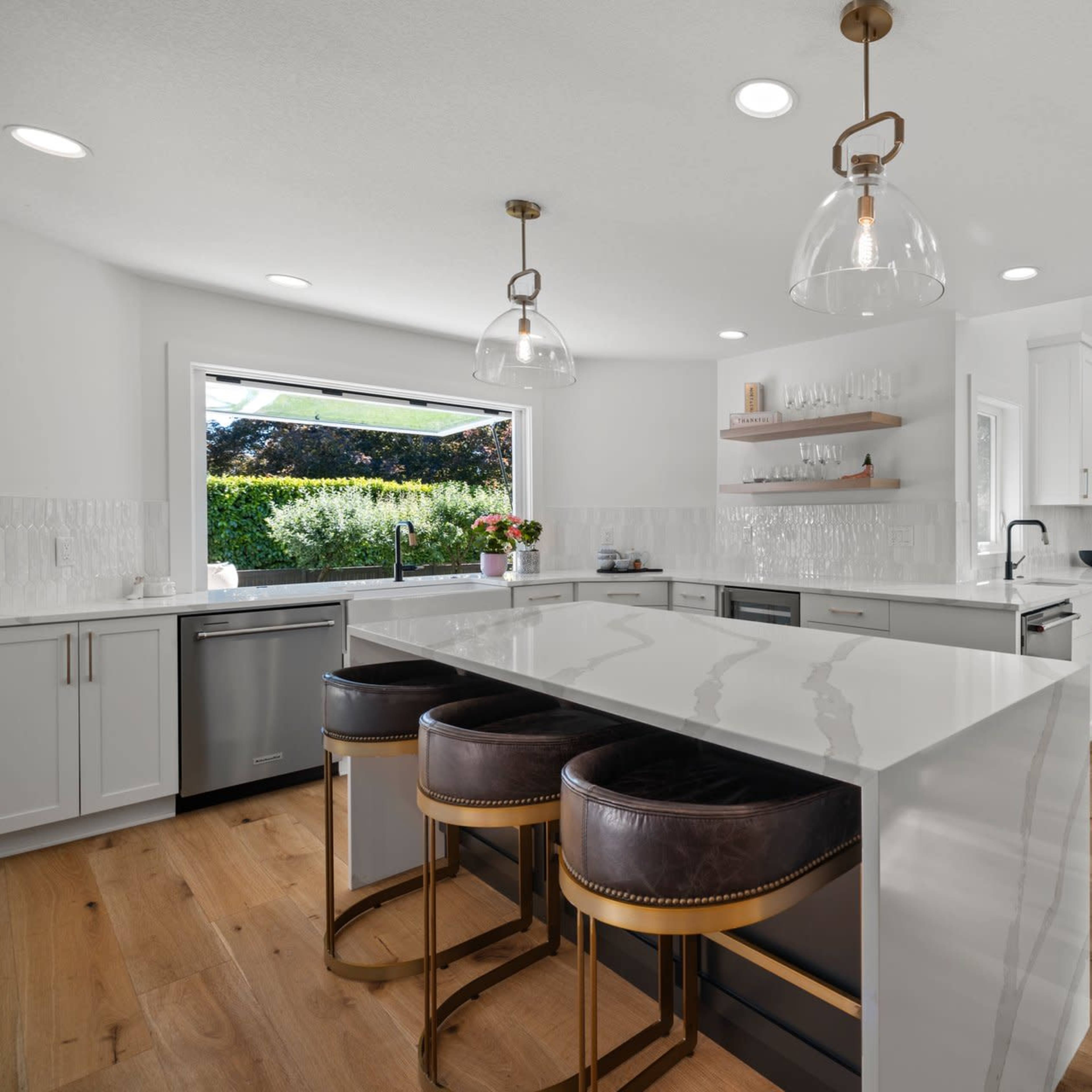 A modern kitchen features a spacious island with three stools, stainless steel appliances, and large windows overlooking a garden.
