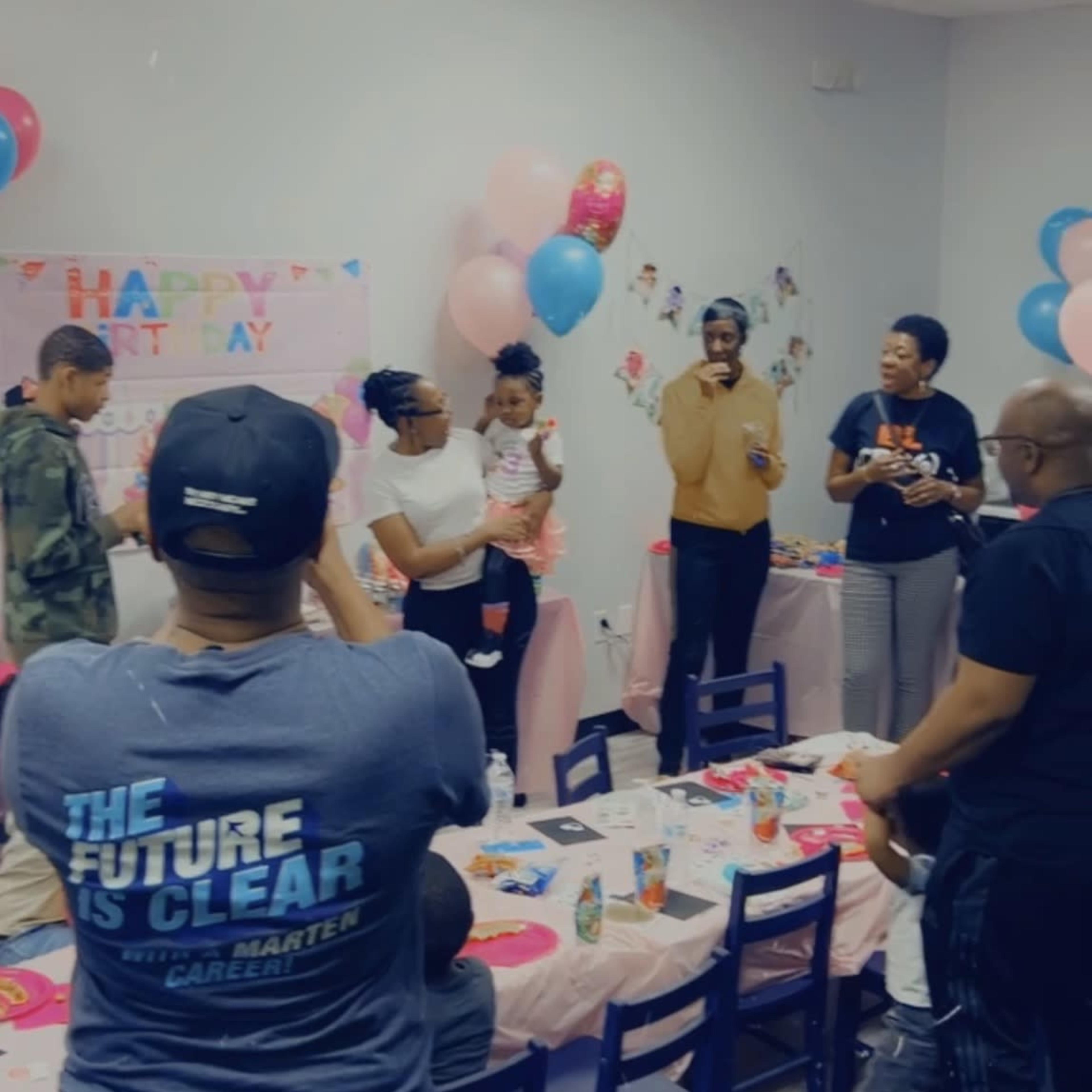 A group of people celebrate a birthday party in a decorated room filled with balloons and a festive table setting.