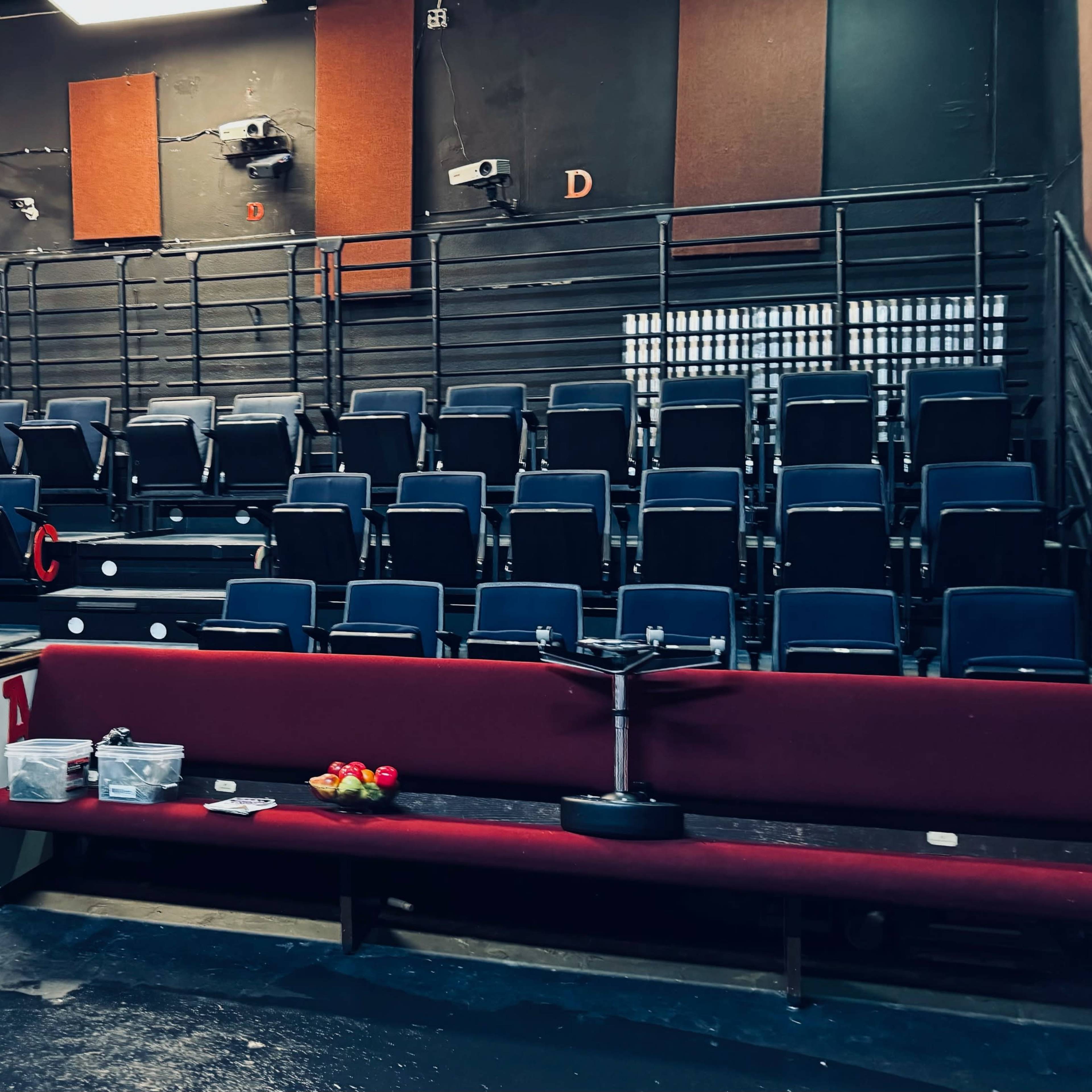 A seating area in a theater features dark blue chairs arranged in rows with maroon benches in front, along with some boxes and items on the ground.