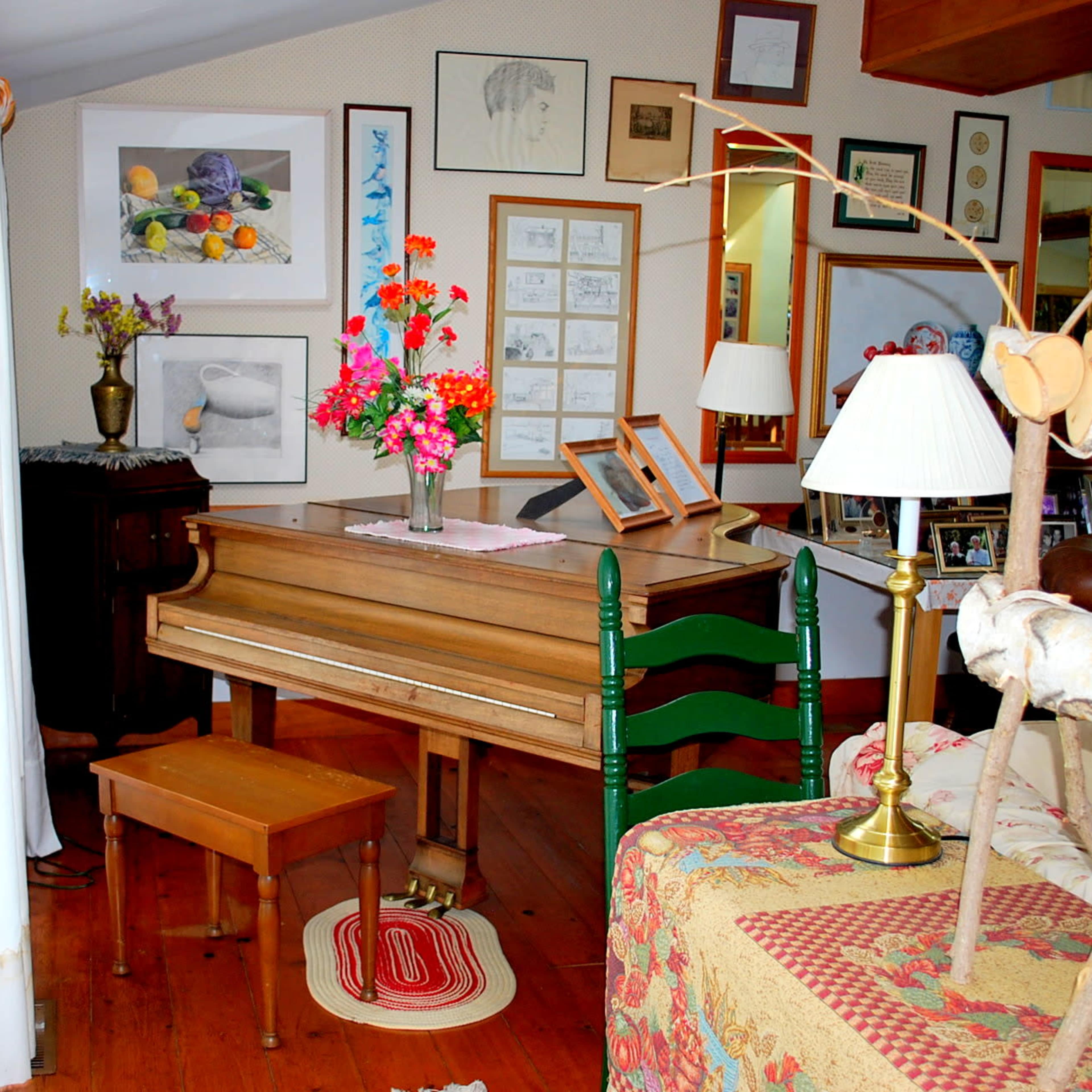 A wooden piano with a matching stool is positioned next to a colorful arrangement of flowers and various framed artworks in a well-lit room.