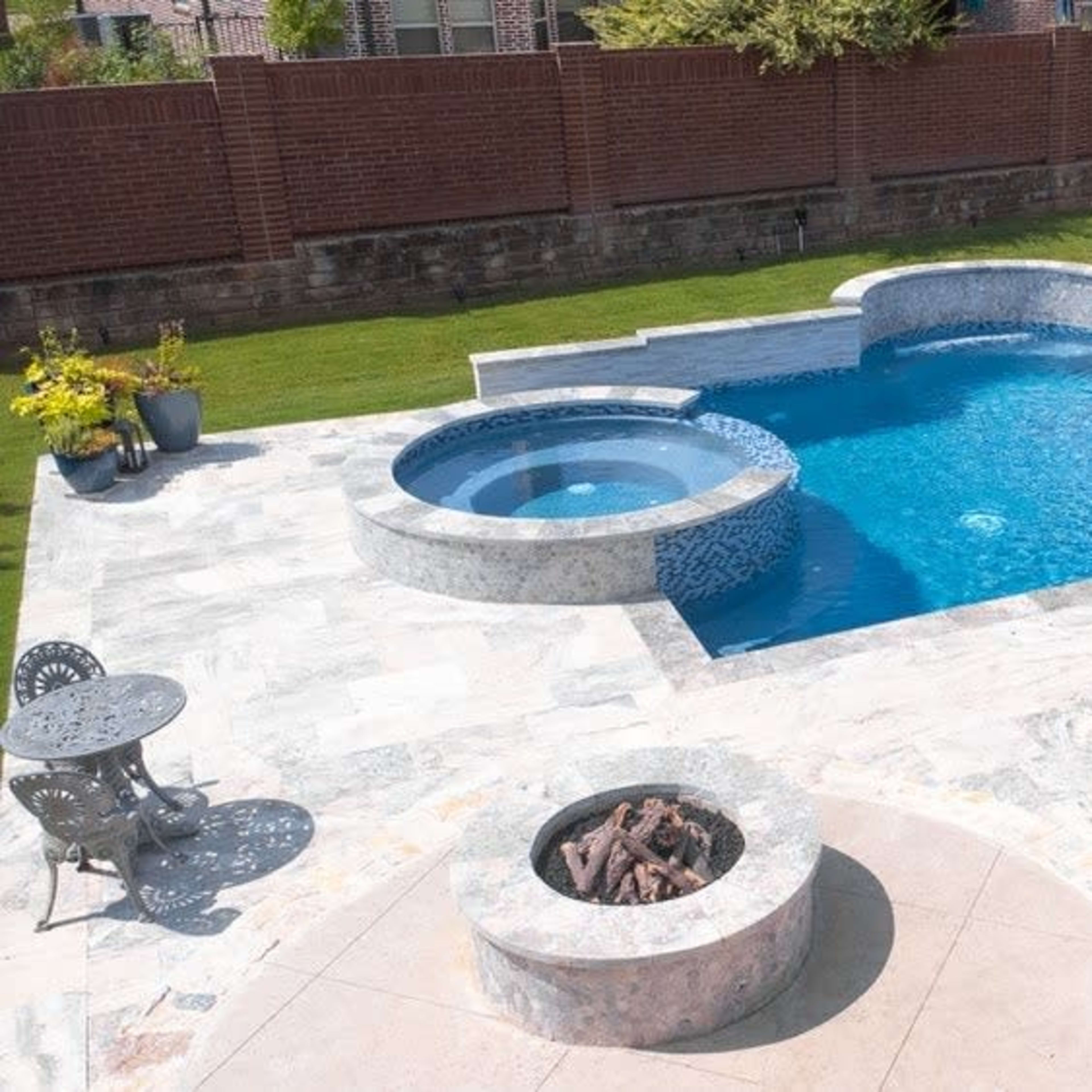 A backyard with a swimming pool, a hot tub, white lounge chairs, and a small fire pit surrounded by stone pavement.