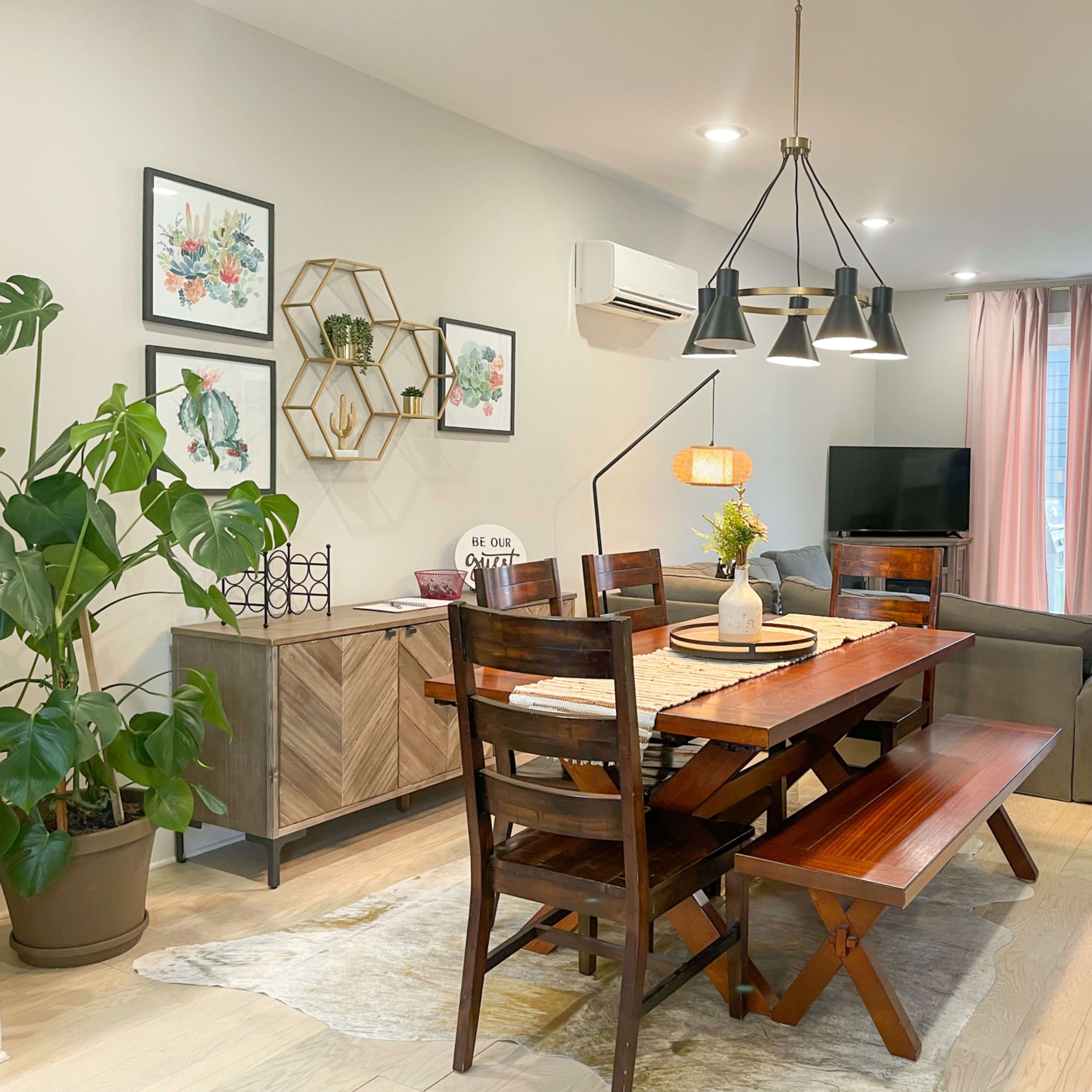 A dining area with a wooden table and benches, surrounded by decorative plants, artwork on the walls, and a living area with a couch and television in the background.