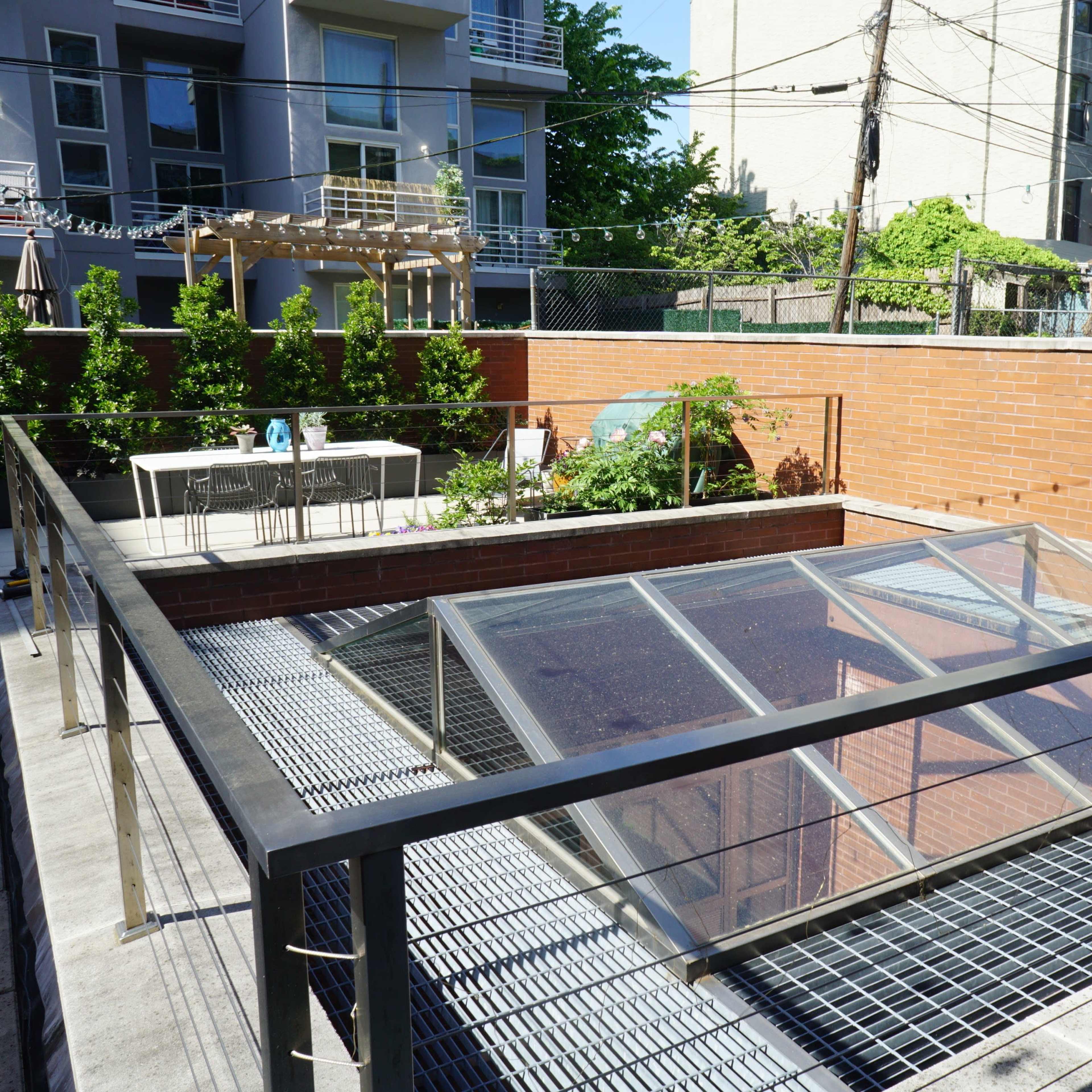 A modern rooftop terrace featuring a glass skylight, dining furniture, and surrounding greenery within an urban setting.