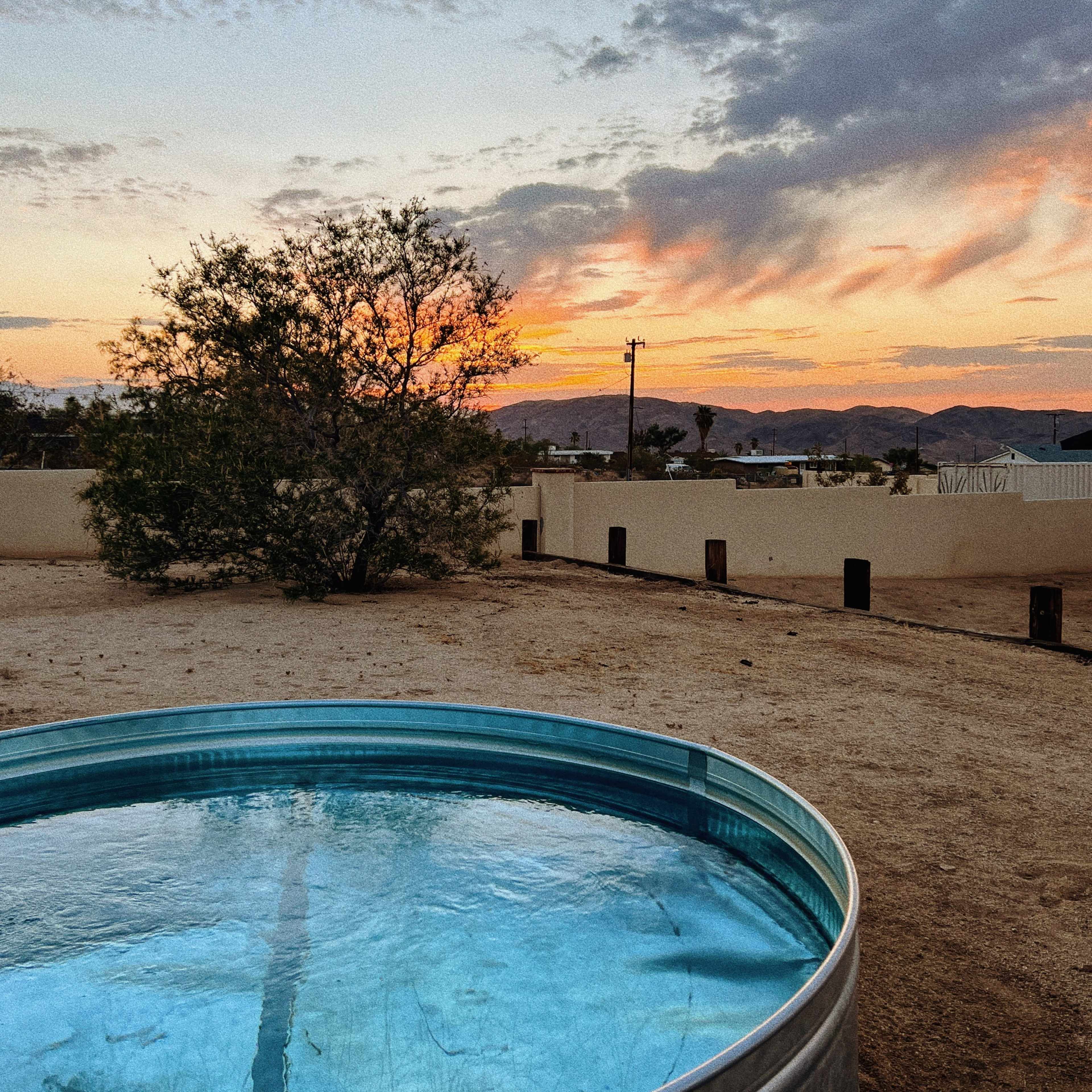A circular swimming pool reflects the colors of a sunset in a desert landscape, with a tree and distant hills visible in the background.