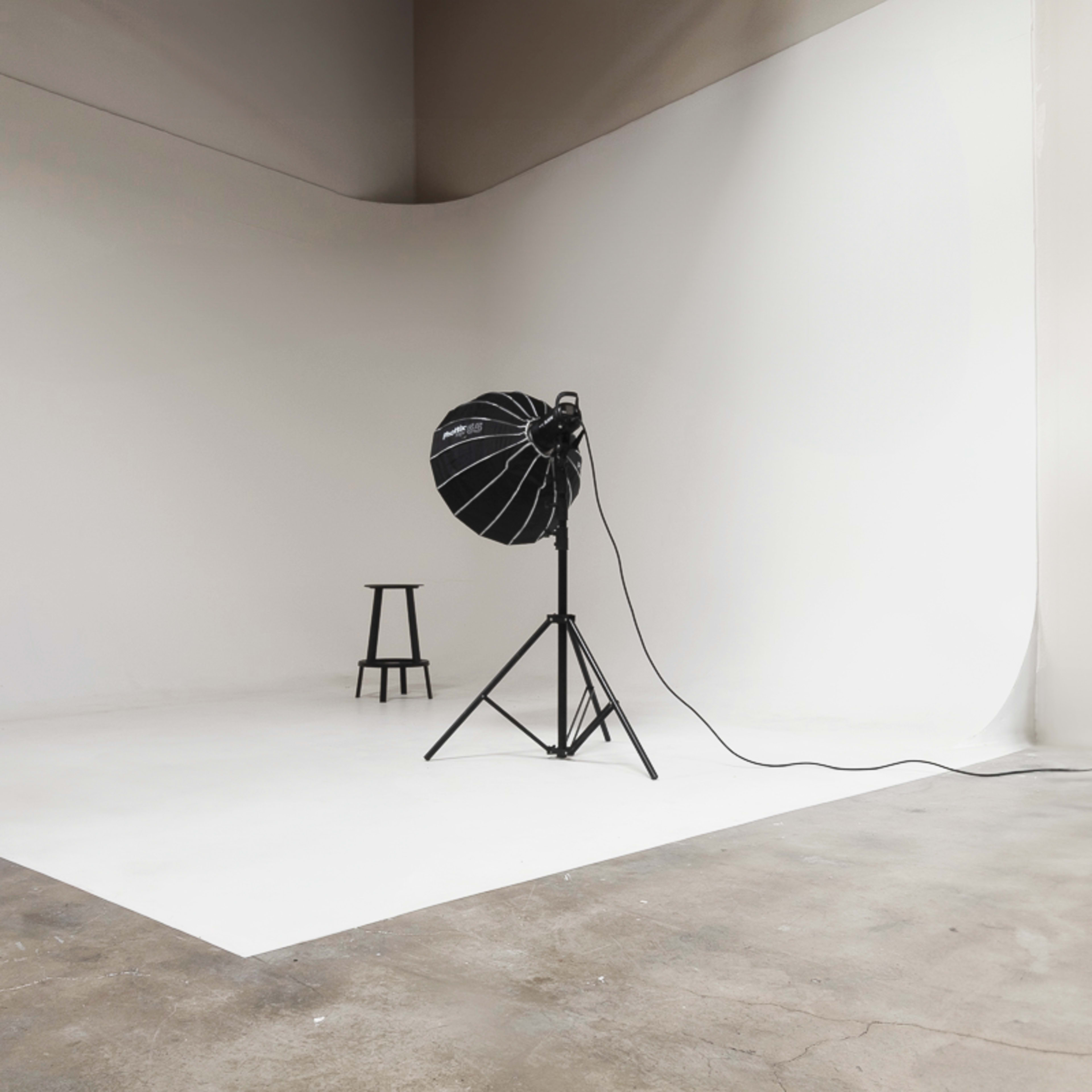 A photography studio features a single black light on a stand and a wooden stool against a white backdrop.