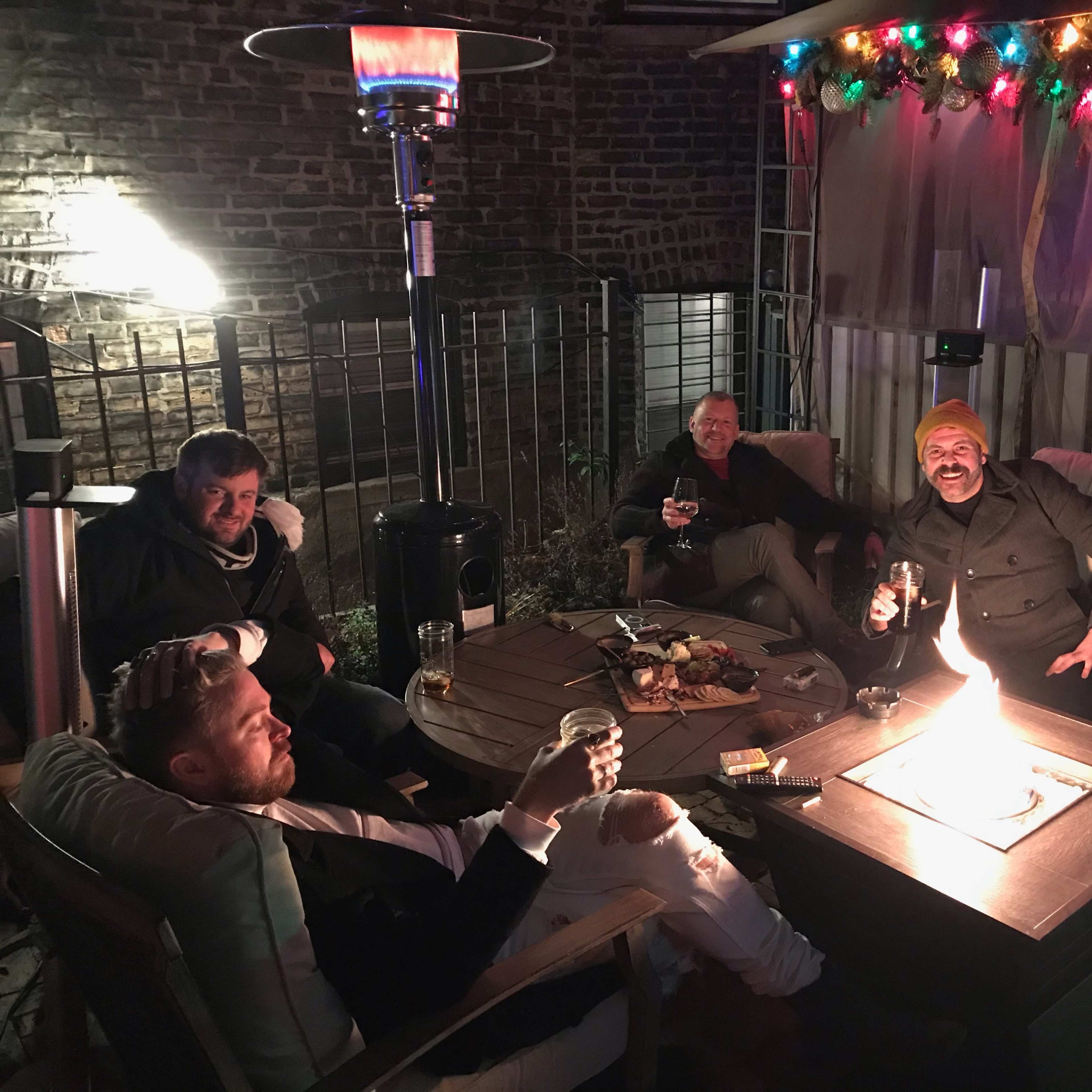A group of five people sits around a lit fire pit on a patio, enjoying drinks and conversation under festive decorations.