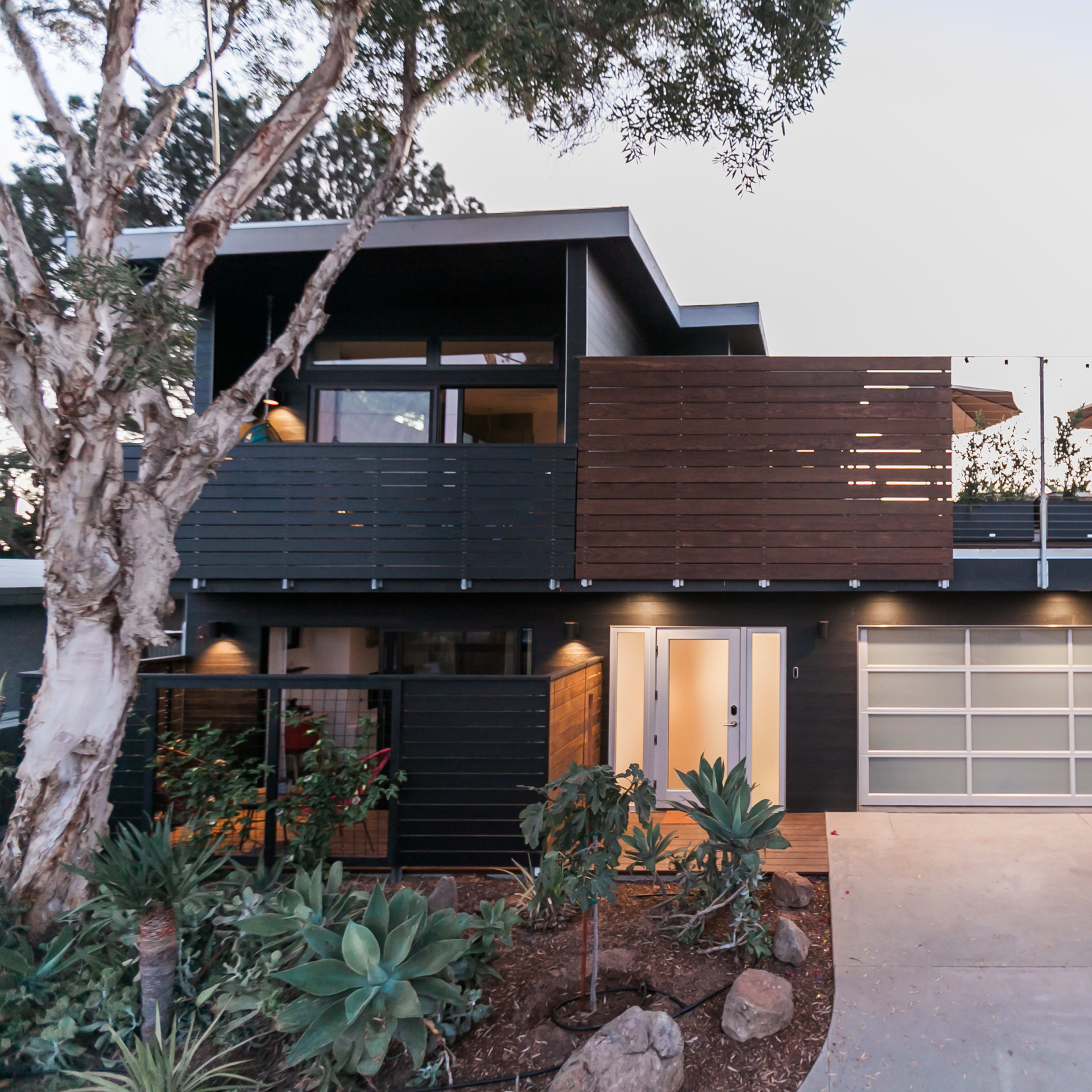 A modern two-story house with dark wood elements, a garage door, and landscaped greenery in front.