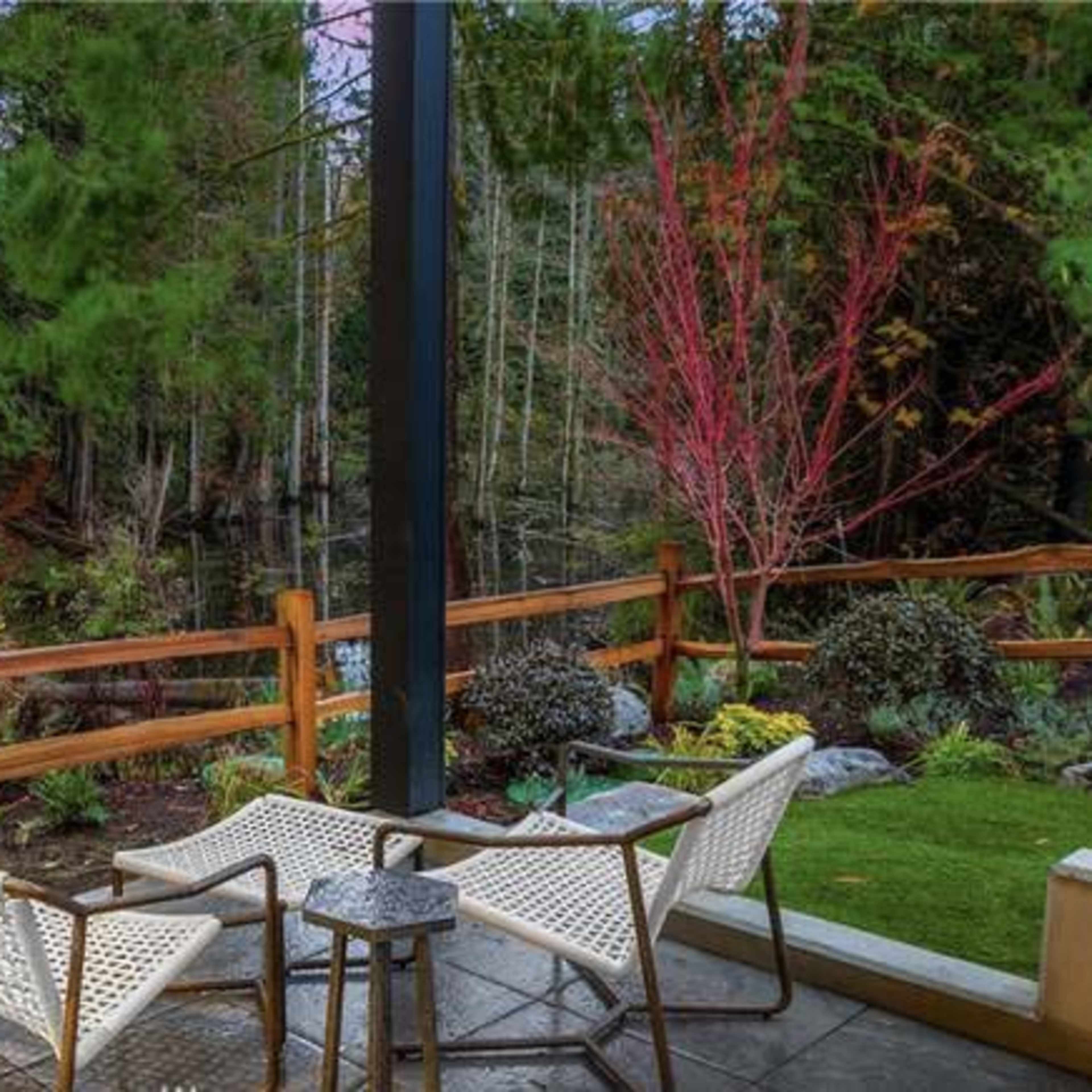 A small outdoor seating area features two chairs and a side table next to a wooden fence, surrounded by lush greenery and trees.