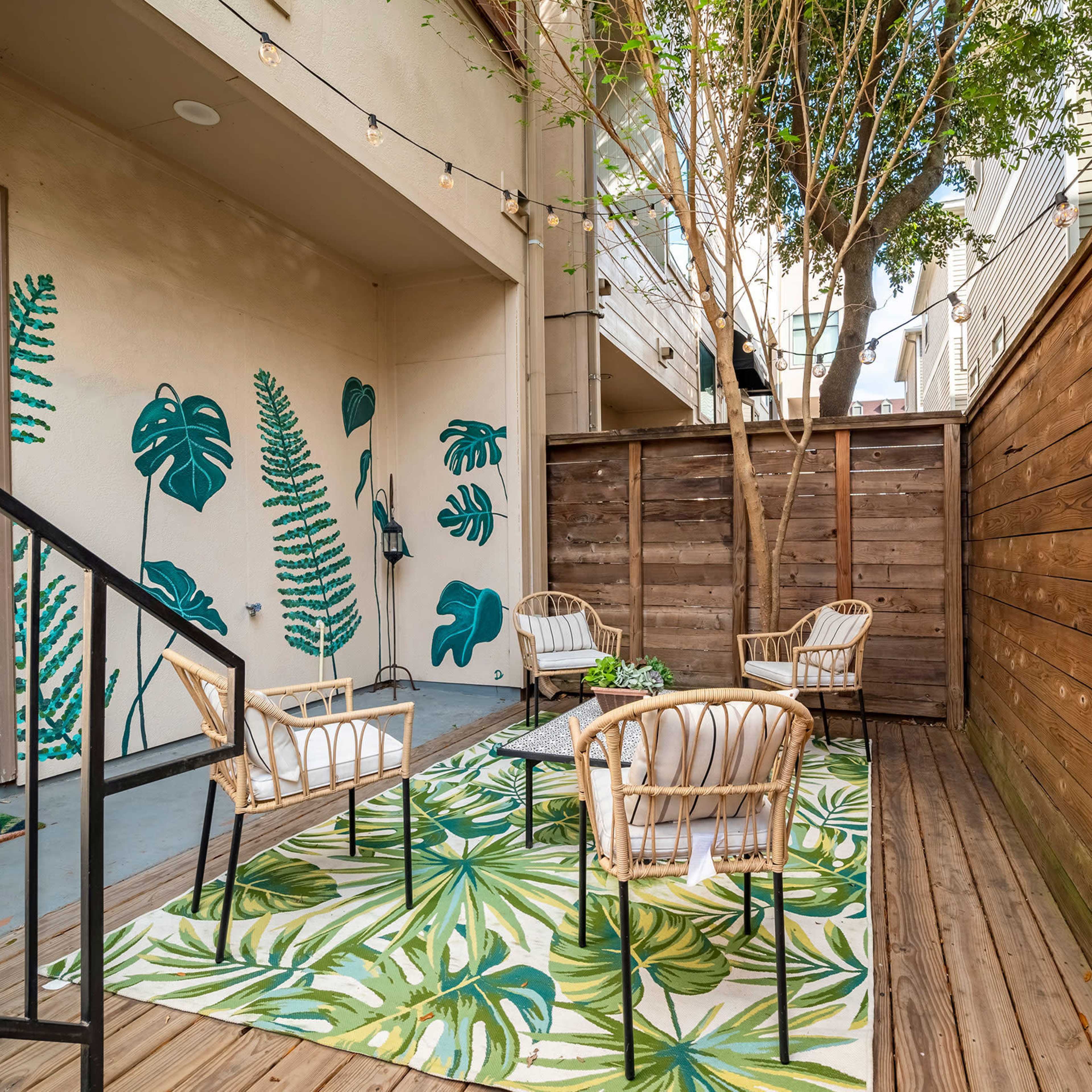 A cozy outdoor seating area features four chairs arranged around a table on a leaf-patterned rug, surrounded by wooden walls and decorative greenery.