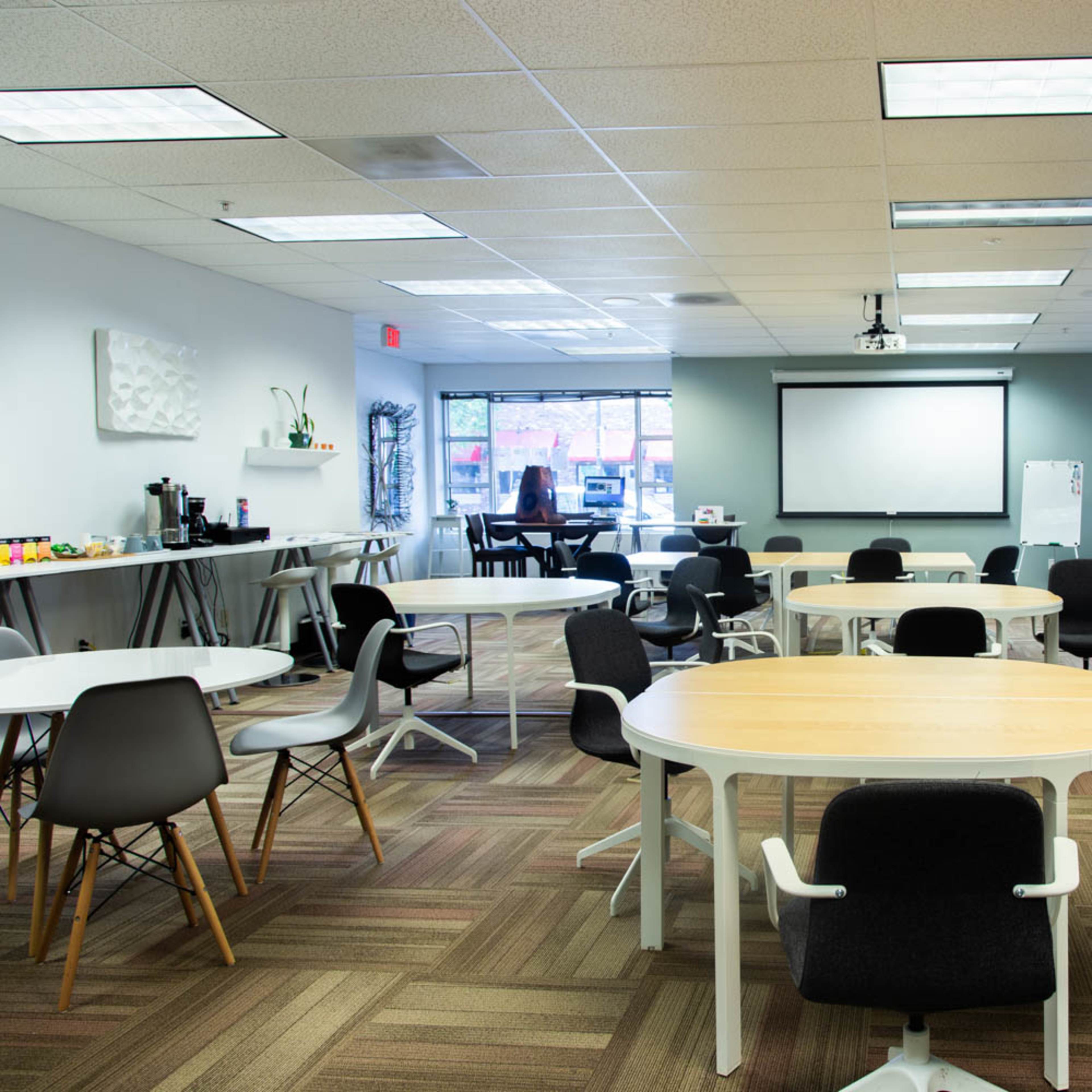 A modern office space with several round and rectangular tables, chairs, and a presentation screen at the front.