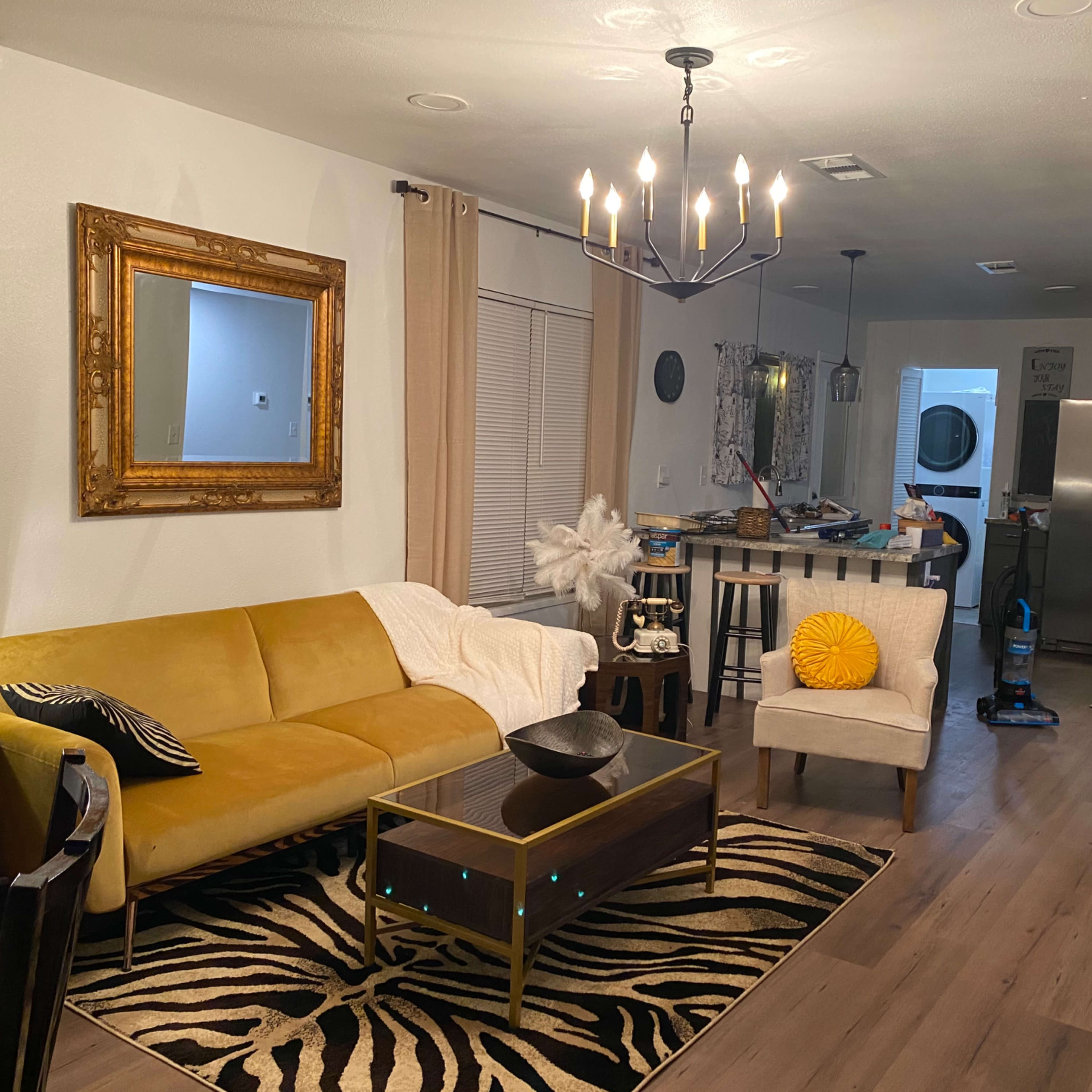 A living room featuring a yellow couch, a coffee table, a white chair with a decorative pillow, and a chandelier overhead, with a kitchen visible in the background.