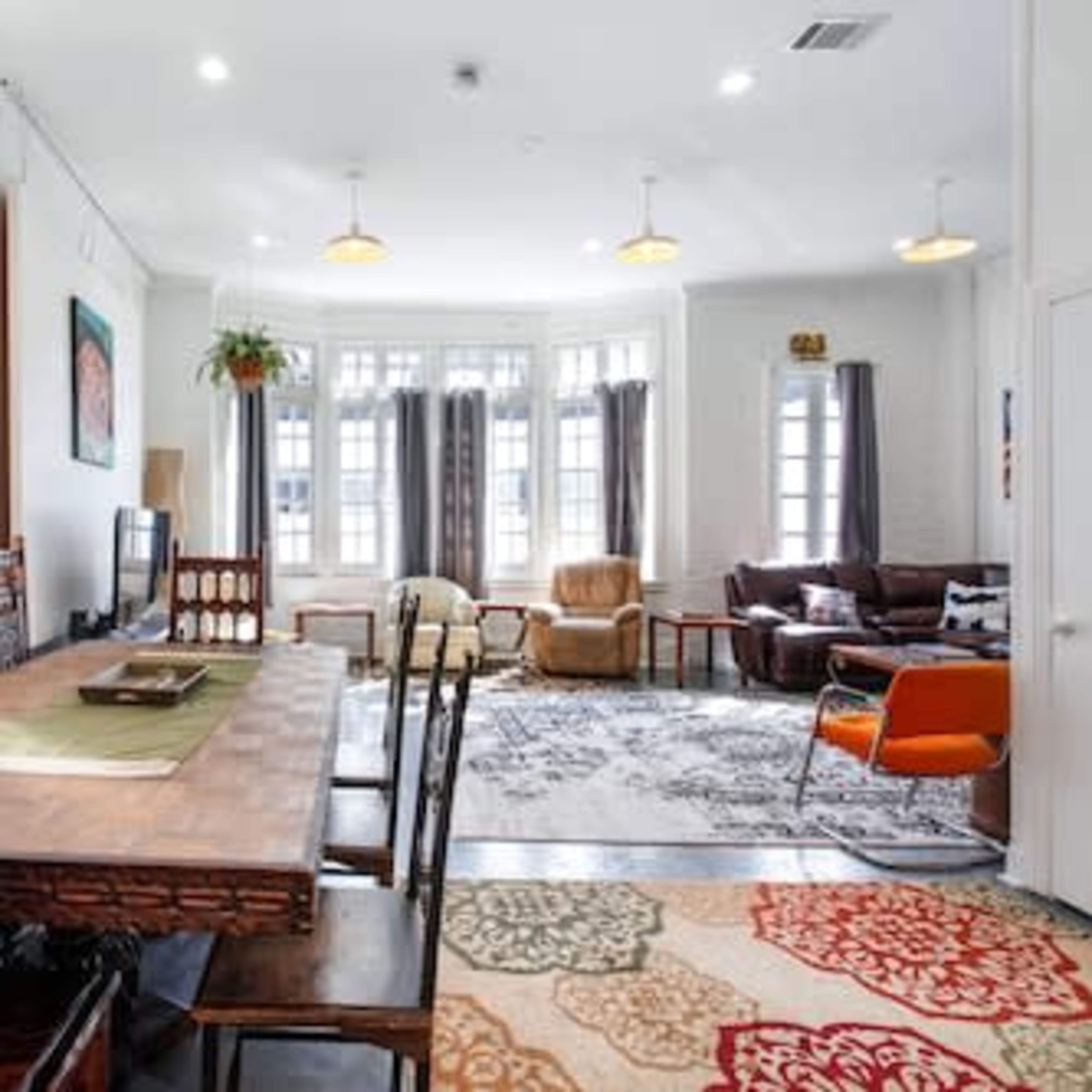 A spacious, well-lit living area featuring a dining table with chairs, a television, a cozy seating arrangement, and large windows.