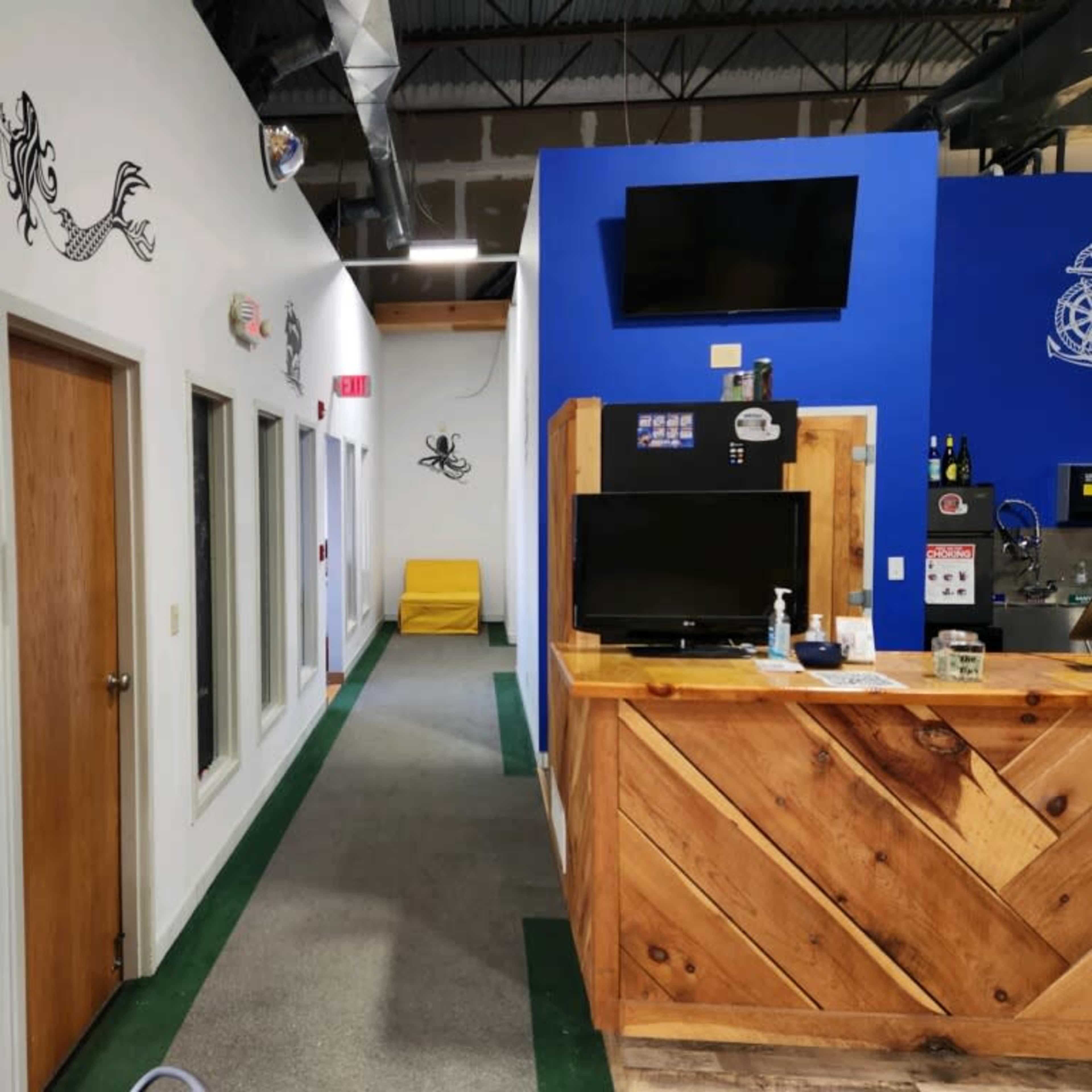 A well-lit indoor space with a wooden reception area on the right, a hallway with doors on the left, and a blue wall featuring a television and decor.